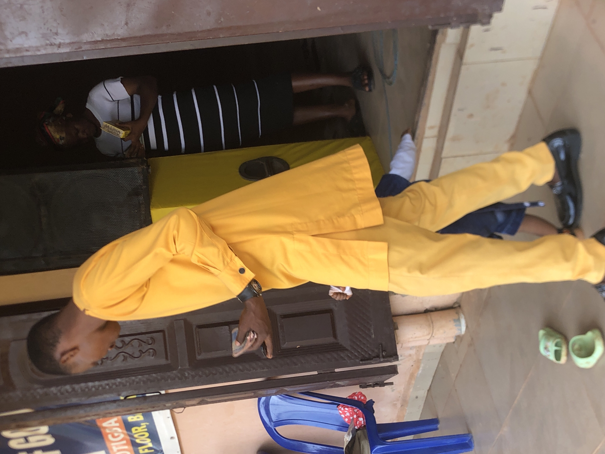 The scene depicts a person in a bright yellow suit standing outside a building in Nkwelle, Nigeria. The person is holding a smartphone. Inside the building, another person wearing a white t-shirt with black vertical stripes is holding a yellow object. Several items are scattered around the foreground, including a blue plastic chair, children's green sandals, and a pipe.