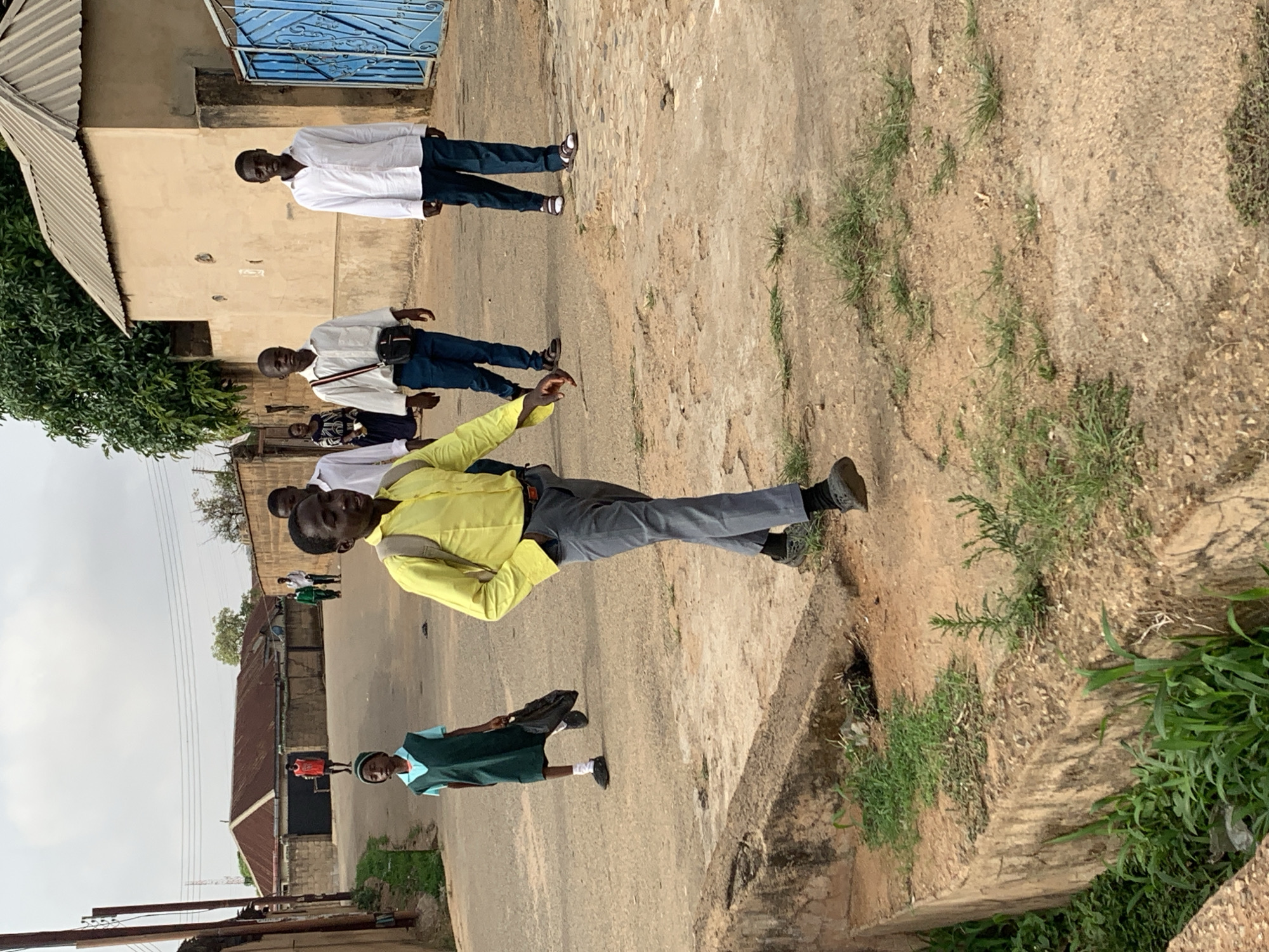 The image depicts a street scene in Jos, Nigeria. A group of people are walking on the street. The foreground includes a concrete curb or edge with vegetation growing beside it. Several people are walking together, with a child in a green uniform, and adults in casual clothing. There are buildings and infrastructure along the street, including metal roofs, power lines, and walls, indicating a populated, urban or semi-urban environment. The sky is overcast.