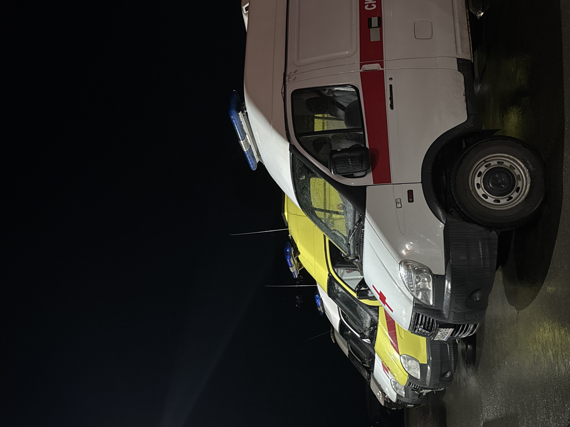 The image depicts a row of parked ambulances in Chaikovsky, Russia. The vehicles are primarily white with red stripes and blue emergency lights. They appear to be lined up on a wet, paved surface at night. The background is dark, with minimal visibility. The focus is on the vehicles themselves, highlighting their details, such as tires, headlights, and body markings.