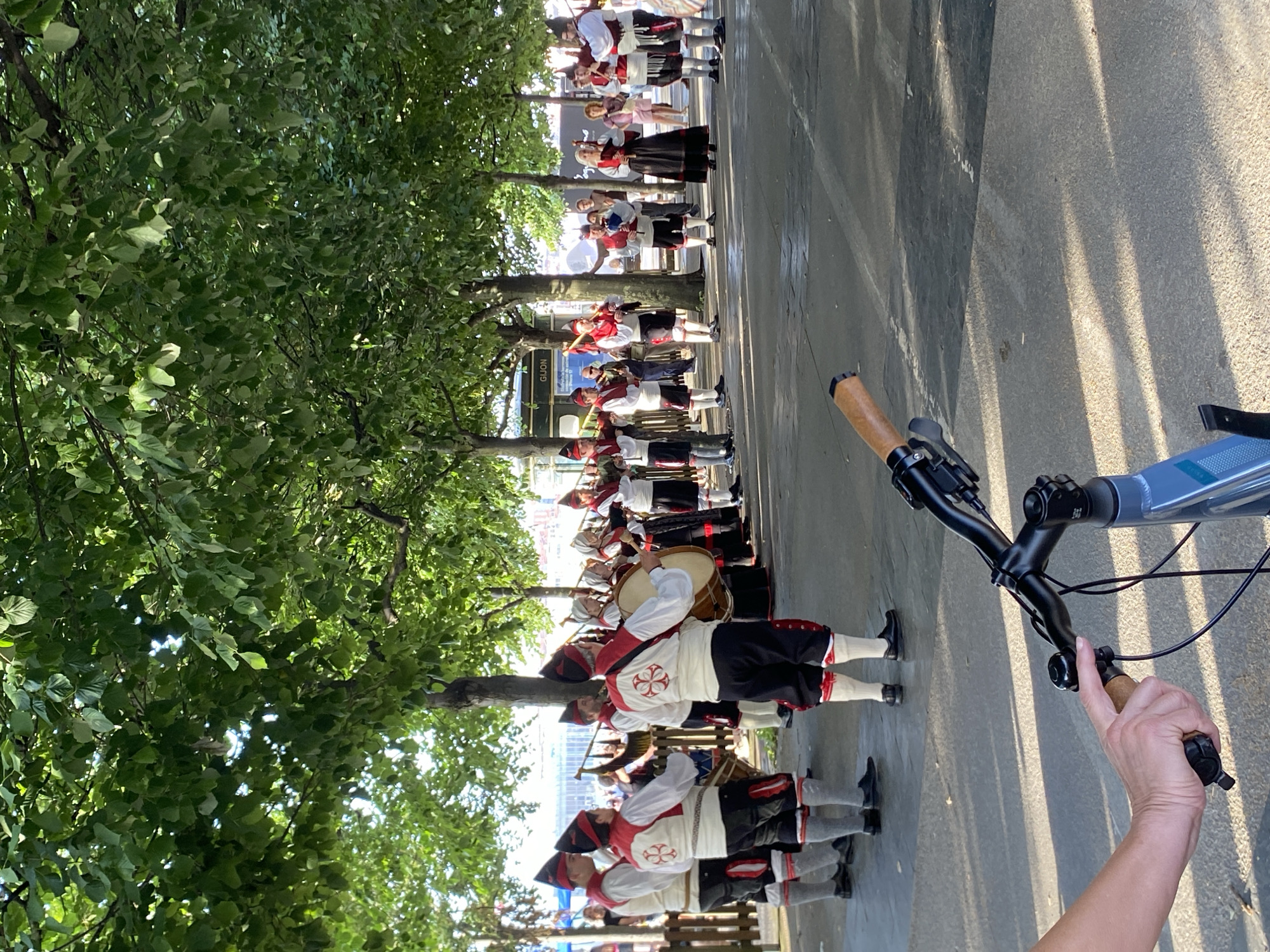 This media file shows a group of people in traditional attire, seemingly participating in a parade or performance. They are lined up along a paved street with trees overhead, suggesting an outdoor event. The individuals are dressed in white shirts with red vests and black pants, some wearing hats. Many are carrying musical instruments, including drums and what appear to be pipes or flutes, indicating a musical procession.
The perspective of the shot is from behind a bicycle, with the handlebars and a person's hand on the grip visible in the foreground. The sun appears to be casting long shadows, suggesting it might be late afternoon or early morning. The overall atmosphere seems festive and orderly, with the participants focused on their activity.
A sign with the text "GIJON" is partially visible on a building in the background, providing a strong location cue that this event is taking place in Gijón, Spain. The weather appears to be clear and sunny, contributing to the outdoor setting.