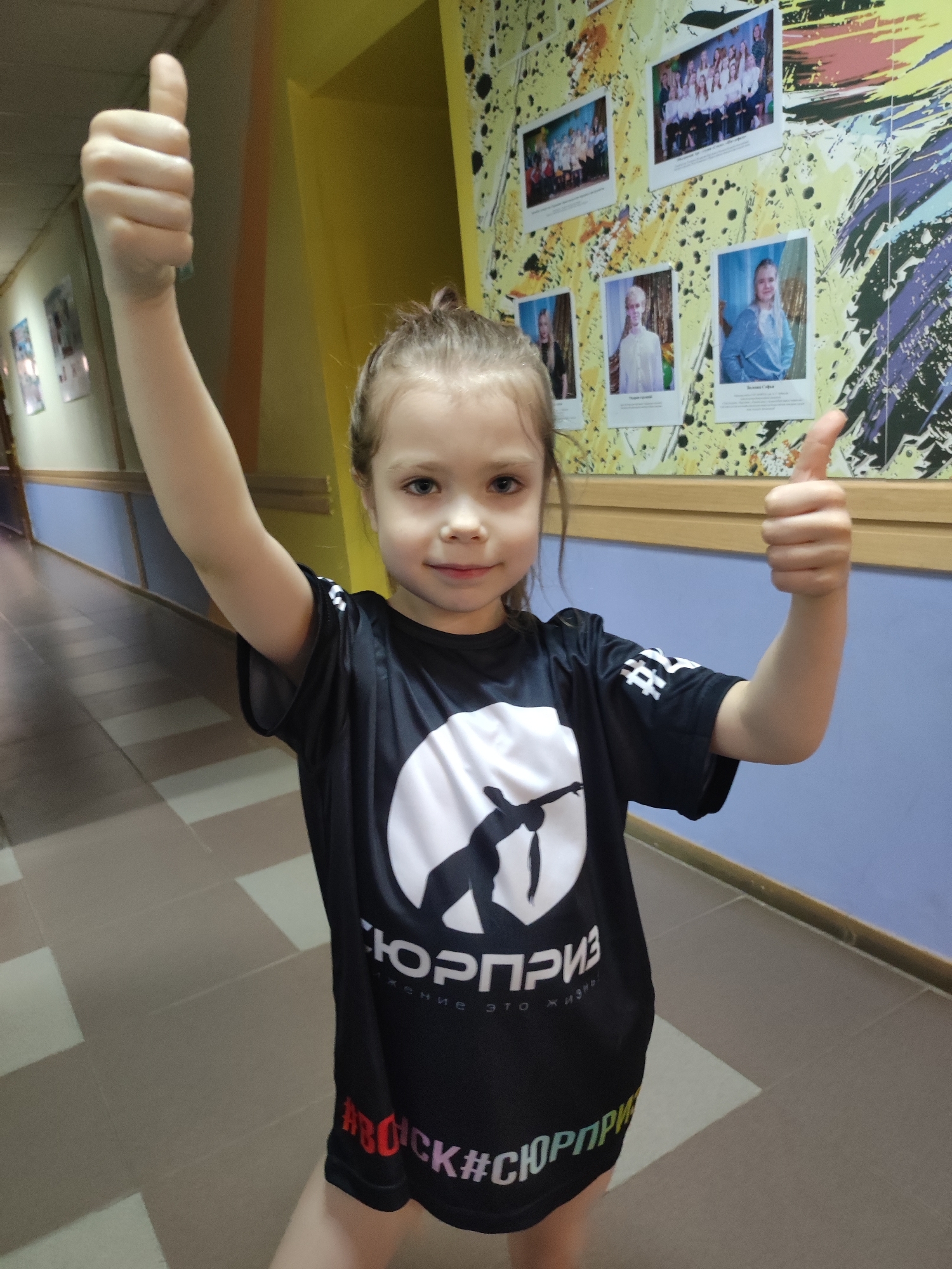 A young girl is standing in a hallway, giving two thumbs up to the camera. She is wearing a black athletic shirt with a white logo and text in Russian. The hallway has yellow walls above a blue section and a gray tiled floor. On the wall behind her are several photographs. The location is likely inside a building, given the hallway. The location may be Votkinsk, Russia.
