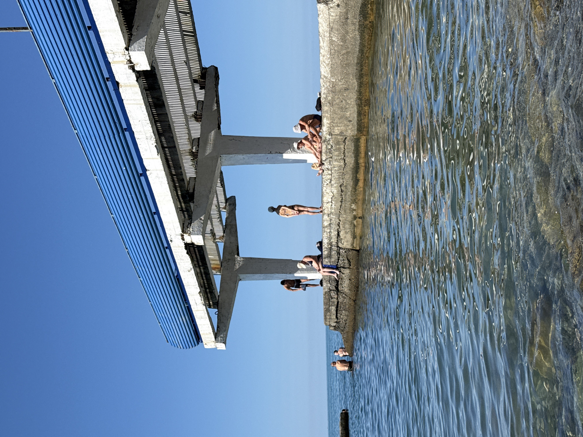 The image depicts a sunny day at a seaside location, possibly in Sochi, Russia, with a concrete pier extending into clear blue water. A blue metal roof structure is supported by concrete pillars over the pier. Several people are present: a few are in the shallow water, while others are sitting or standing on the pier, some appearing to be sunbathing. The water is calm with some gentle ripples. The sky is a clear, bright blue. The concrete structure appears to be somewhat aged, with visible supports and some weathering.