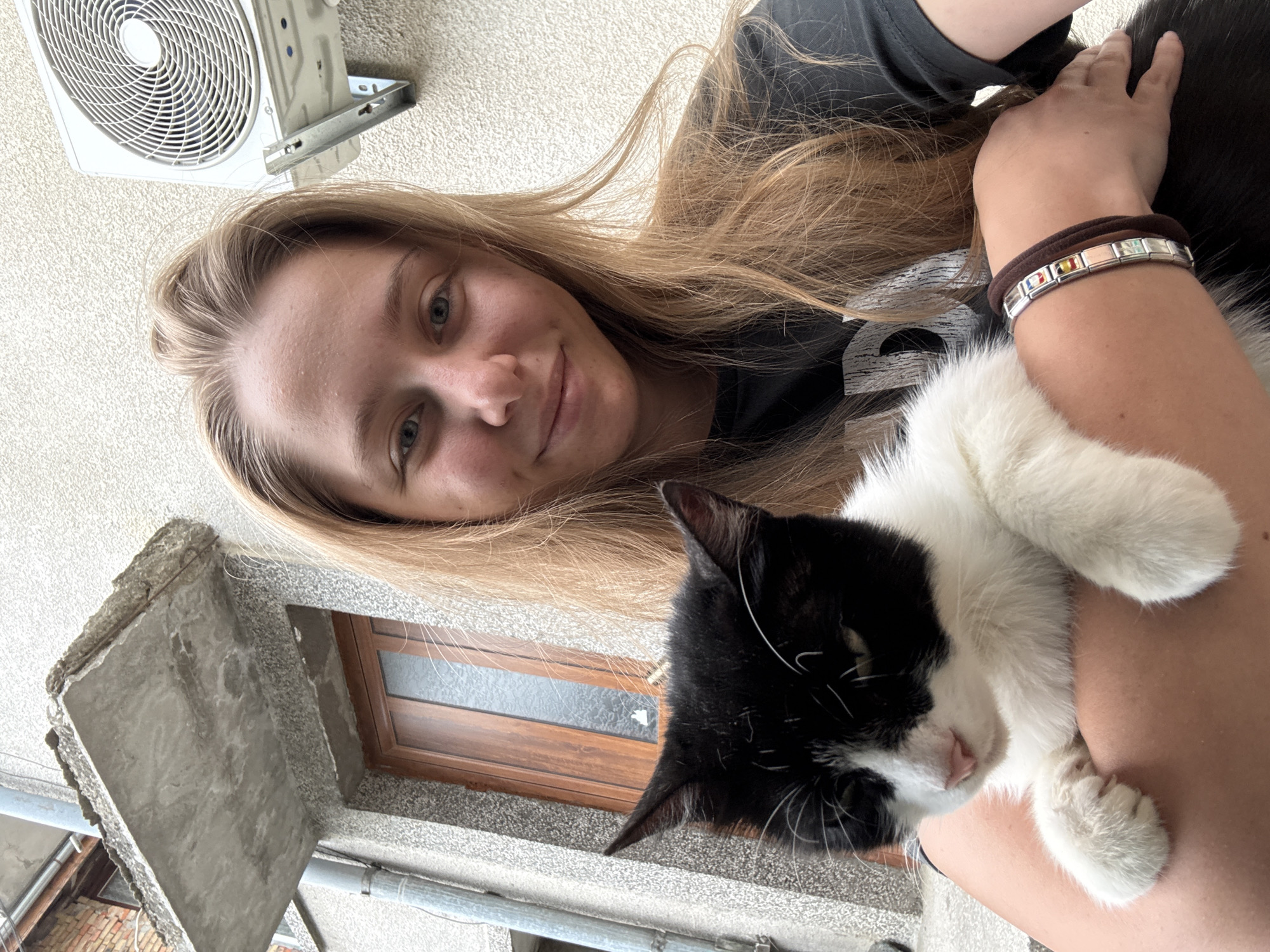 A young woman with long, blonde hair is holding a black and white cat in her arms. She is wearing a black t-shirt and a bracelet on her left wrist. The cat is looking down. They are outdoors in front of a building with a textured, light-colored wall. An air conditioning unit is mounted on the wall to the right of the woman. To the left of the woman, a section of a weathered concrete overhang is visible above a wooden door. The image appears to have been taken in Tsarevo, Bulgaria.