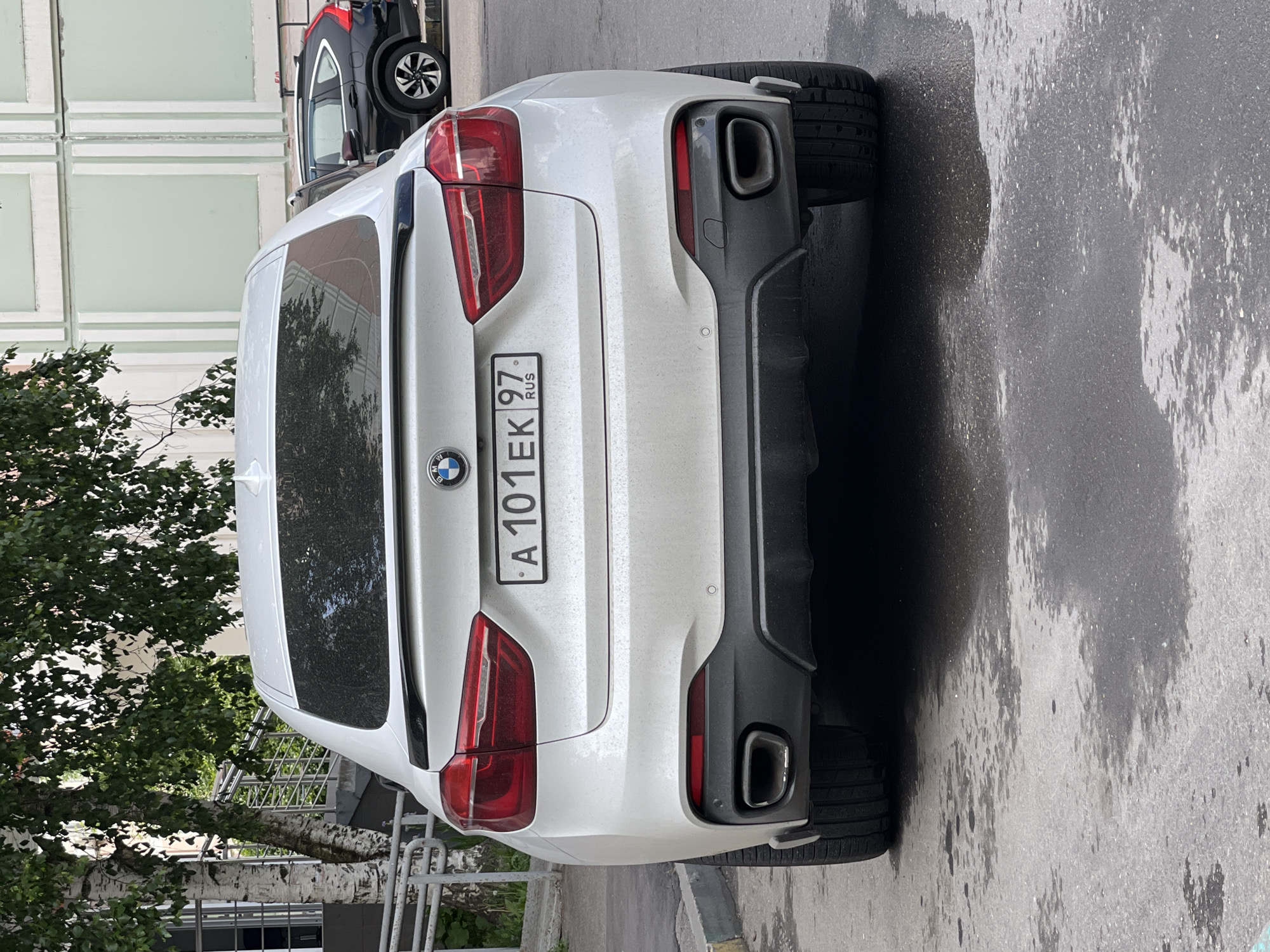 The image shows the rear of a white BMW X6 parked on an asphalt surface, presumably a street or parking lot. The car has a Russian license plate that reads "A 101 EK 97 RUS." The surroundings include a building with a light green facade and window grid and a tree with green leaves. The location is likely Moscow, Russia, based on the "RUS" marking on the license plate. There are no people visible in the image. Another vehicle, partially visible, is parked behind the BMW. The asphalt surface appears slightly wet.