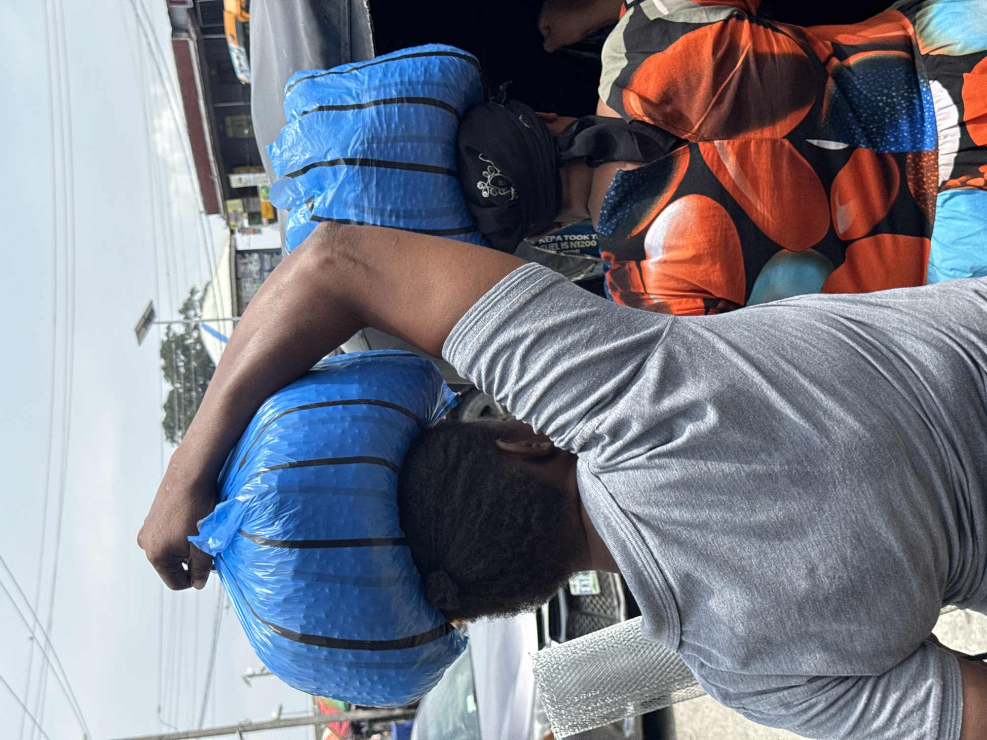 This is a medium shot photograph taken from behind two people in a city environment, likely Ado, Nigeria, based on the provided context. The sky is overcast, suggesting possibly a hazy or cloudy day.
The foreground features the backs of two individuals. The person on the left is wearing a grey, short-sleeved polo shirt. Their right arm is raised, supporting a large, bright blue plastic bag on their head. The bag appears to be tightly packed and has vertical black stripes. Part of a silver, mesh-like object is visible in front of this person's left side.
To the right of the first person, another individual is visible, also with a large, blue plastic bag on their head. This bag also has vertical black stripes and is placed over a dark headscarf adorned with white embroidery. This person is wearing a colorful, patterned garment with large orange and blue shapes on a dark background.
In the background, a street scene is discernible. A silver car is parked or moving to the left of the frame. Further in the distance, buildings with awnings and signs can be seen, and a solar panel is visible on a rooftop. There are also power lines crisscrossing the sky. The overall impression is that of a busy marketplace or street in an urban setting.