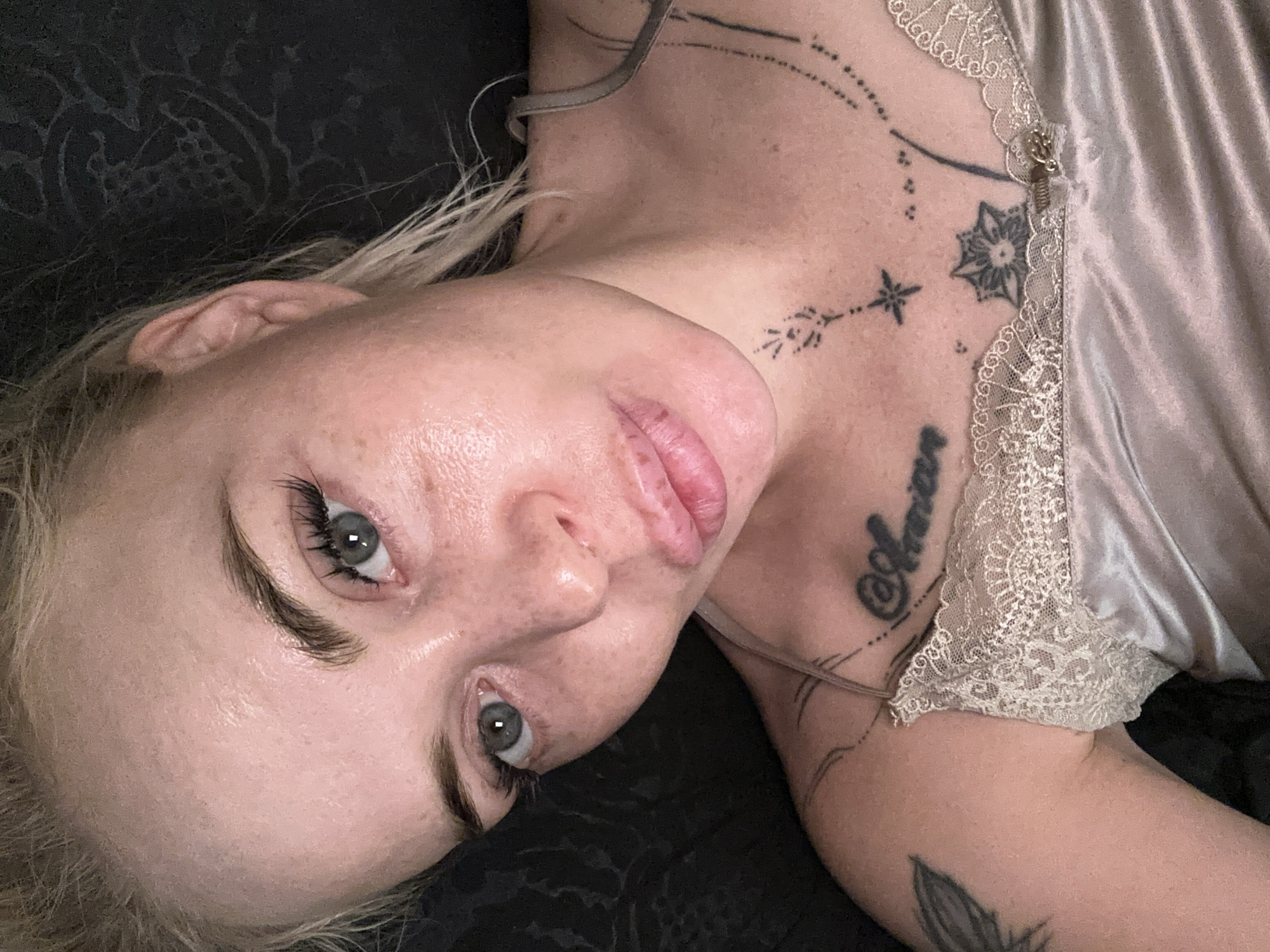 A medium shot of a fair-skinned woman with blonde hair, light green eyes, and freckles is captured from a high angle. Her eyes are looking forward, and her mouth is slightly open. She is wearing a light-colored camisole with lace trim. There are several tattoos visible on her chest and shoulder, including script and floral designs. The background is dark and appears to be a patterned fabric. The lighting is dim, creating a soft, intimate atmosphere. The image was taken in an unknown indoor location.