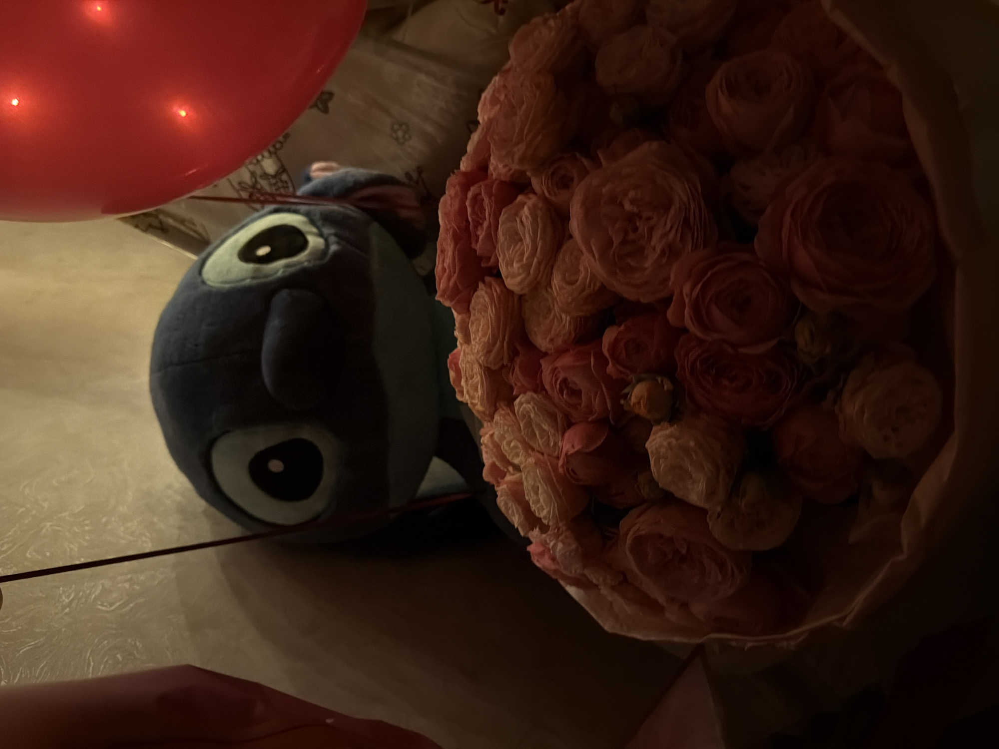 The scene features a red balloon, a plush toy resembling the character Stitch, and a bouquet of pink roses. The balloon is positioned to the left of the frame. The Stitch toy is next to the balloon. The bouquet of roses is arranged to the right, wrapped in paper. The location is Chaikovsky, Russia.