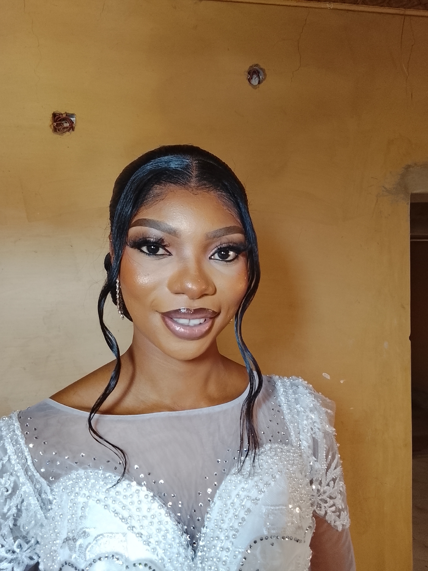 A head and shoulder indoor shot shows a young Black woman with a dark complexion, dark hair styled with two curly strands falling on either side of her face, and wearing a white dress with lace sleeves and rhinestone embellishments. Her makeup is heavy, with dark eyeliner, false eyelashes, and glossy lips. She is facing the camera and smiling, with her head slightly tilted to her left. The background is a textured wall, painted in a muted yellow color. On the wall there are two electrical outlets with loose wiring. The location is in the city of Unknown, Unknown.