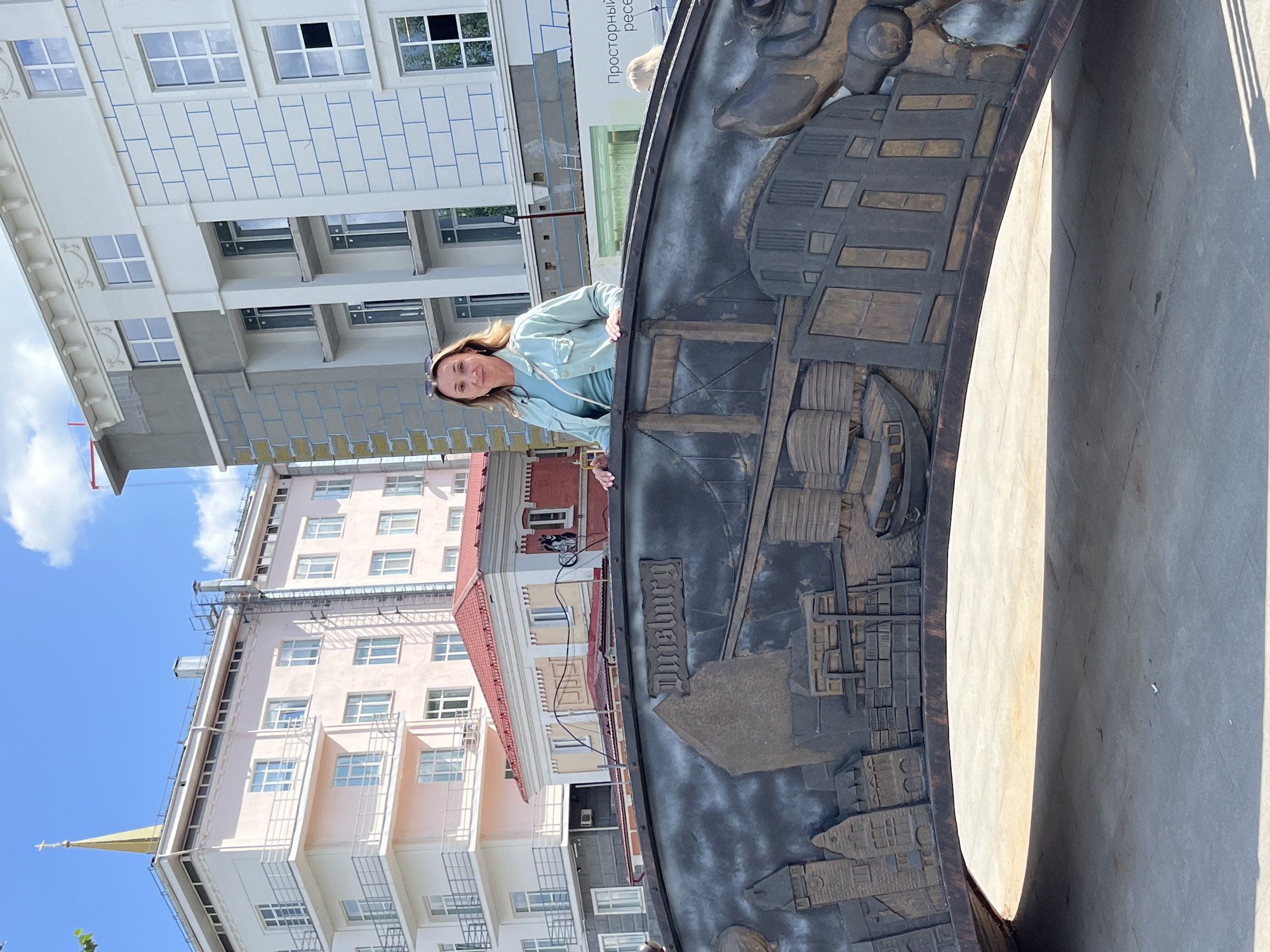 A woman stands behind a decorative bridge railing in the city of Perm, Russia, with buildings visible in the background. The bridge railing is made of dark metal and features a bas-relief depicting buildings and a ship. The woman is wearing a light blue denim jacket over a turquoise t-shirt and sunglasses pushed up on her head. She is smiling and looking at the camera. Behind her, several buildings of varying architectural styles are visible, including a light pink building with a gold spire topped with a cross. Another building to the right is under construction, with visible scaffolding. The sky is blue with scattered clouds, indicating a sunny day. The ground in front of the bridge is paved with grey concrete. The overall scene is an outdoor urban environment.