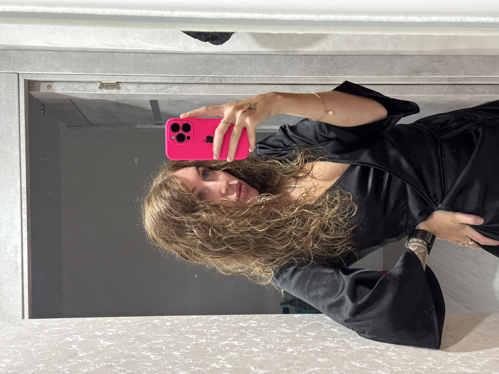 A woman with curly, light brown hair is taking a selfie in a mirror. She is wearing a black, low-cut dress with flared sleeves. She is holding a bright pink iPhone in her right hand. Her left hand is on her hip, and she is wearing a black watch on her left wrist. A gold bracelet is on her right wrist. A light-colored counter and a grey-toned mirrored cabinet are visible in the background. The photograph was taken at a tilted angle. There is no verifiable information to suggest this photo was taken in Chaikovsky, Russia.