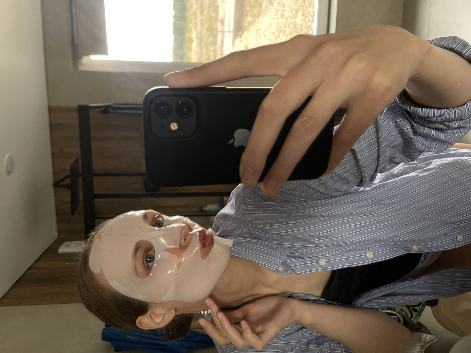 A woman is taking a selfie in what appears to be an indoor setting. She is wearing a light blue and white striped shirt. She has a sheet mask on her face. She is holding a black smartphone with an Apple logo, and she is holding a gold necklace. The location is not discernible to be the city of Подшивалово, Russia.
