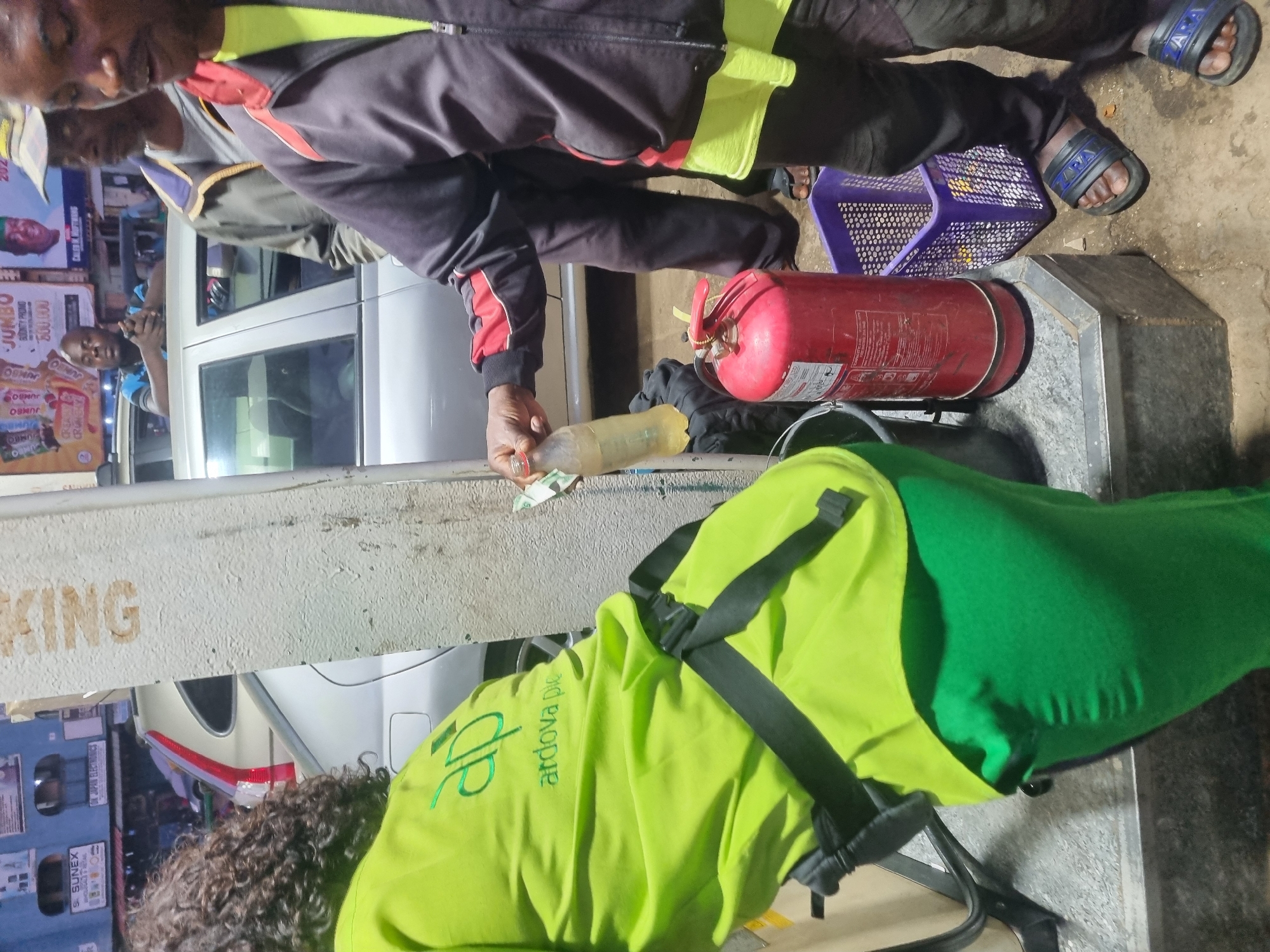This image shows a nighttime scene in what appears to be Jos, Nigeria. A man wearing a black and red jacket and sandals is pouring liquid from a yellow plastic bottle onto some money. Nearby, there's a red fire extinguisher and a purple plastic basket on a concrete surface. Another person wearing a green and yellow vest with the words "ardova pic" is also present. In the background, a car is visible along with a sign containing the word "KING" and a poster advertising a "JUMBO BONUTY PROMO" for 500,000.