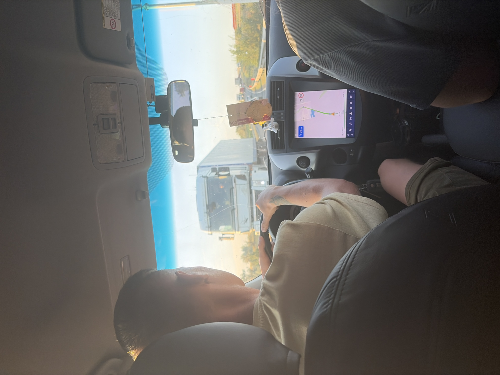 An image taken from inside a car shows a man driving on a highway. He is wearing a light-colored t-shirt and has his hands on the steering wheel. A large white truck is visible in front of the car. The dashboard displays a navigation system with a map. A decorative air freshener hangs from the rearview mirror. The environment appears to be a sunny day with a clear sky. The location is identified as Belogorki, Russia.