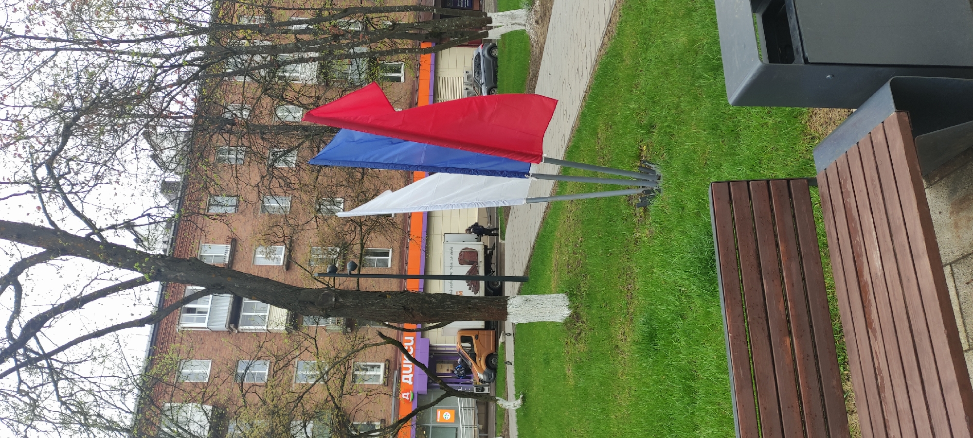The photograph, taken in the city of Fryazino, Russia, shows a street scene with red, white, and blue flags standing in a grassy area adjacent to a brick building. A tree is situated near the flags, and a wooden bench sits in the foreground. A sidewalk runs alongside the grass, and parked vehicles are visible in the background. A person is seen near a truck in the background, and a commercial building is present with signage in Russian. The weather appears overcast.