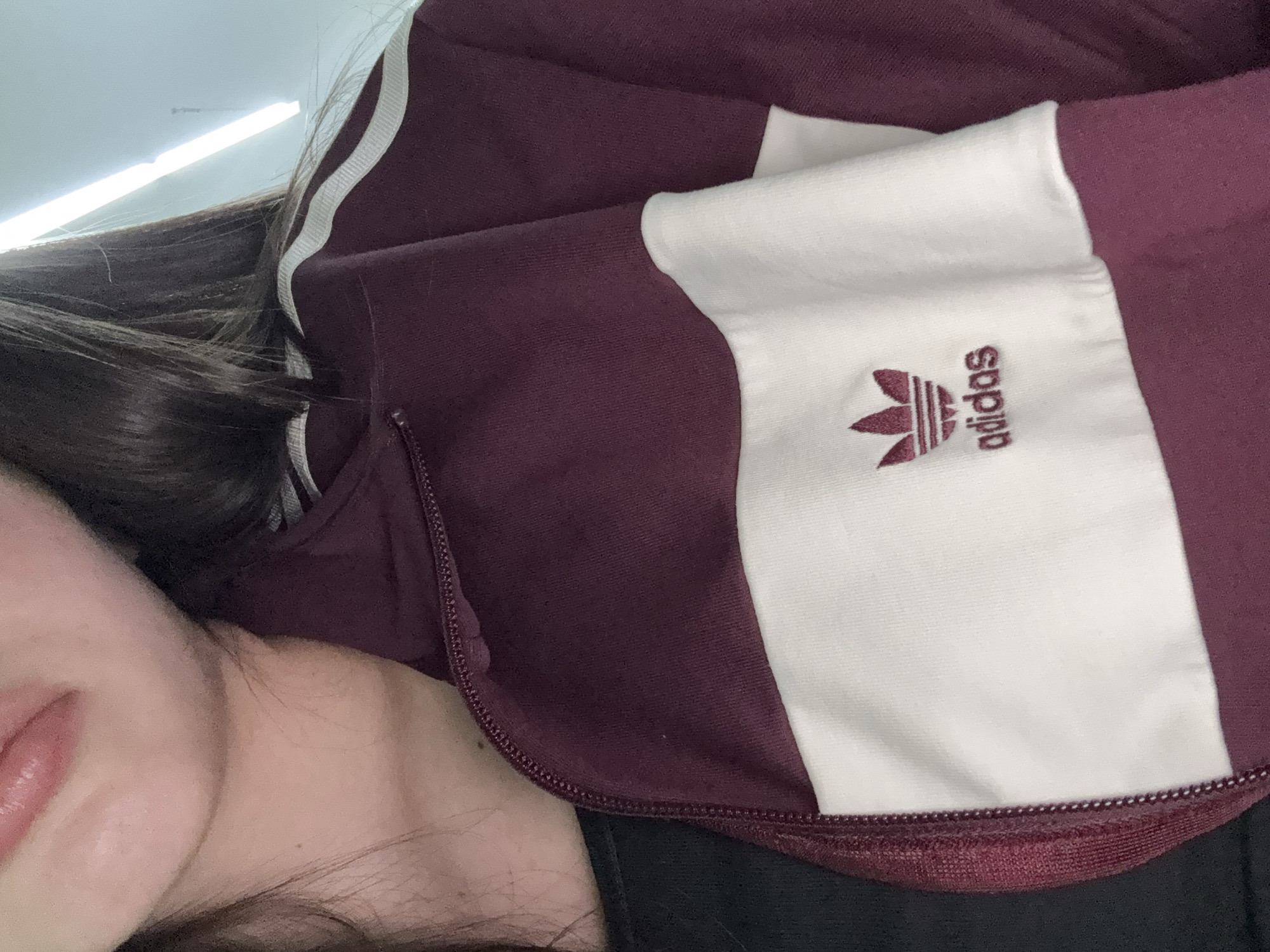 The image shows a partial view of a person wearing a burgundy Adidas jacket with white accents and the Adidas logo. The jacket is zipped up. The person's face is partially visible, showing their lips and chin. The environment is not discernible enough to place it in a specific location in Yerevan, Armenia, or any other location. The lighting appears to be from an overhead source, possibly fluorescent. The person is wearing a black shirt under the jacket.
