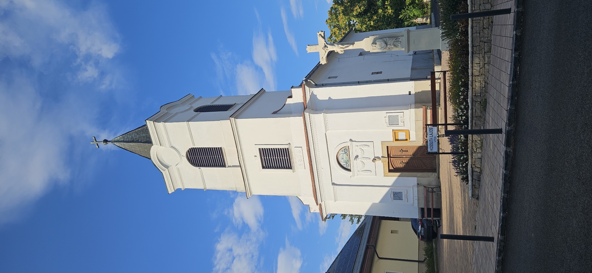 The image shows the exterior of a white church with a dark, peaked steeple topped with a cross. The church has arched windows with louvers and a date, "1833," inscribed above the entrance. A large wooden door with a cross design is central to the facade. To the right of the church entrance is a statue of Jesus on the cross with another smaller figure at its base, dated "1898." There are two wooden benches outside the church and a sign that reads "KOSSUTH LAJOS UTCA." A parked car is visible to the left of the church, alongside a building with solar panels. The scene is set against a bright blue sky with scattered clouds. The location is identified as Balatongyörök, Hungary.