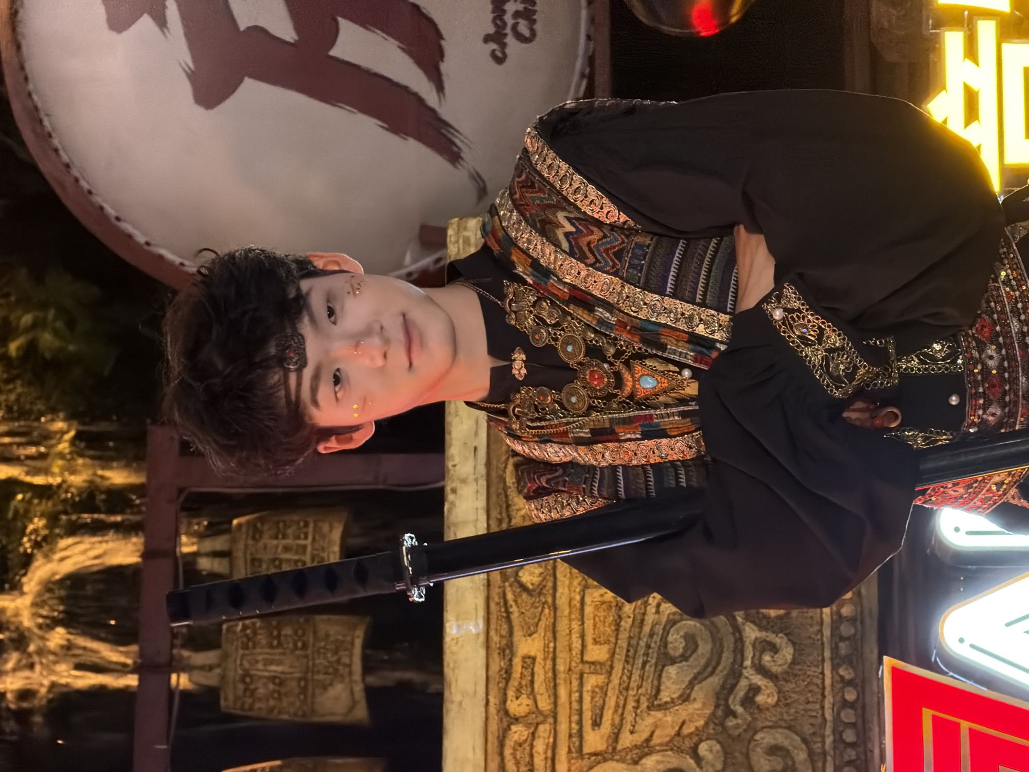 A young East Asian male stands in front of a blurry background that appears to be outdoors at night. He is wearing a black, long-sleeved outfit with a high collar and ornate gold and multi-colored fabric accents on the shoulders and sleeves. He also has multiple layers of gold necklaces and decorative chains around his neck. A black sword with a silver hilt is held vertically in front of him, with his arms crossed over it. He has dark, curly hair and is wearing a decorative headband. His face is turned towards the camera, and he has a slight smile. There are gold specks of glitter on his cheeks and nose. In the background, a large drum with a red symbol is visible, along with what appear to be traditional Chinese bells and glowing neon lights. The overall impression is one of a cultural performance or festival. The image is likely taken in Yuzhong District, China, given the context of traditional Chinese cultural elements.