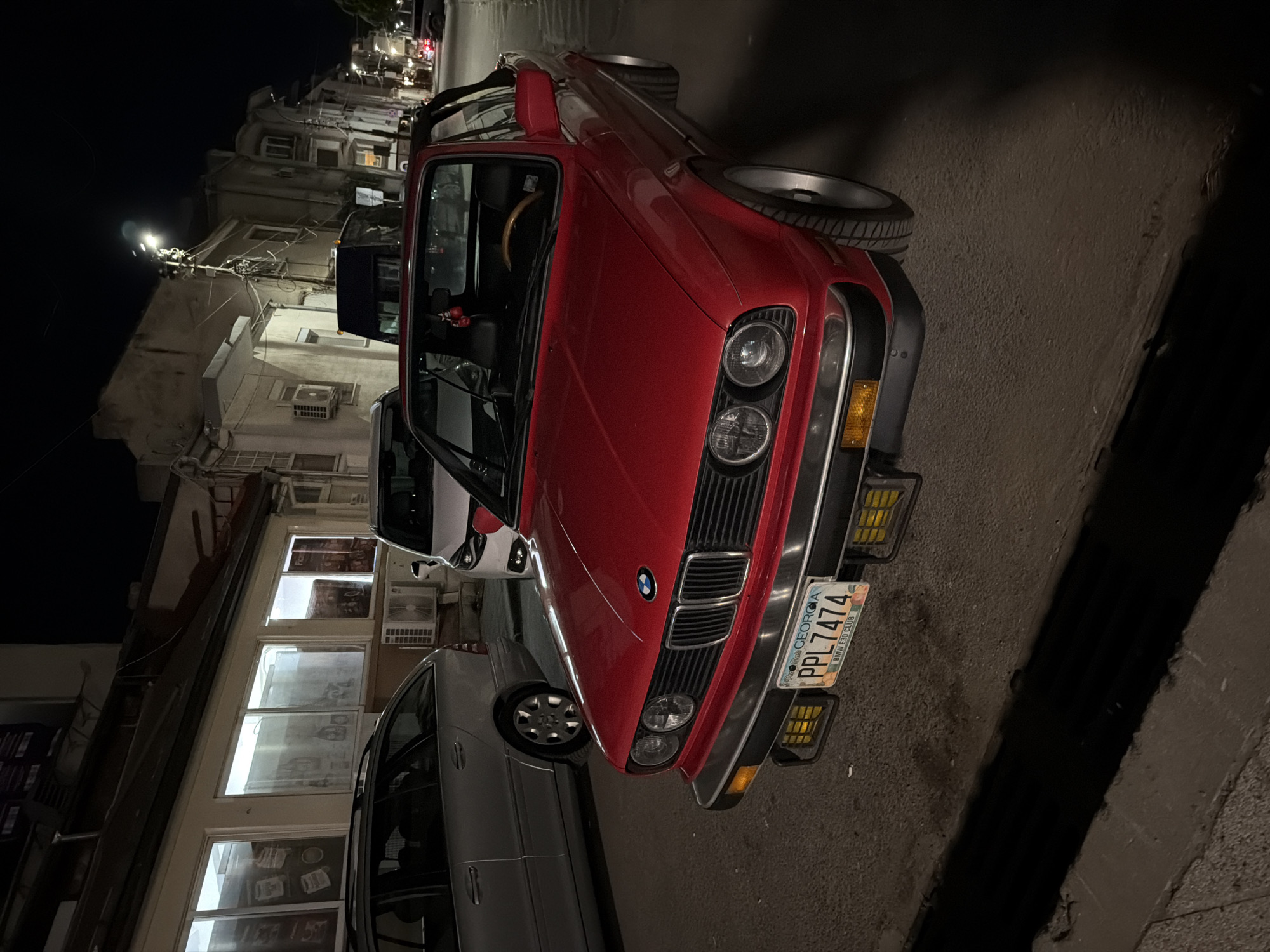 The image shows a red BMW E30 convertible parked on a street at night, likely in Burgas, Bulgaria. The car is the primary subject, facing slightly to the right, with its front visible. It has a Georgia license plate reading "PPL7474". To the left of the red car is a silver station wagon. Further back, a white van is parked. The background reveals buildings with lit windows and streetlights, indicative of an urban environment. The scene is illuminated by artificial light sources, creating shadows and a nighttime atmosphere. There are no people visible in the image. The context of Burgas, Bulgaria, is an external detail not directly derivable from the visual content of the image itself.
