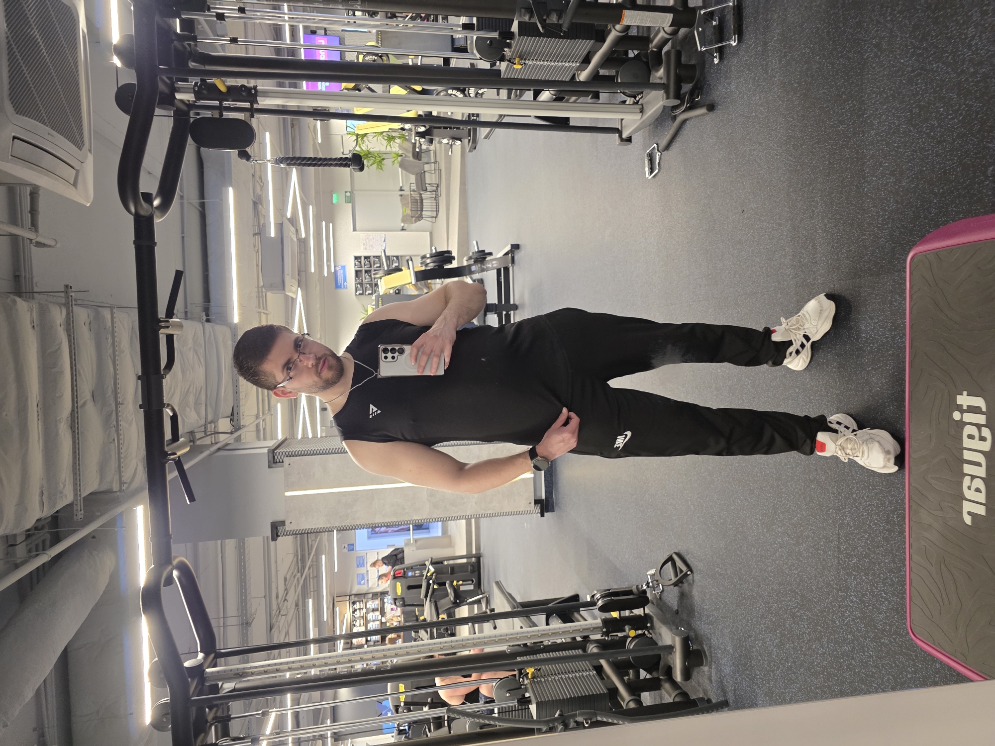 A man in a black tank top and black Nike sweatpants is standing in a gym. He is holding a phone and taking a selfie. He is wearing glasses and a watch. The gym has exercise machines and equipment. The location is not explicitly specified but is understood to be in Plovdiv, Bulgaria.