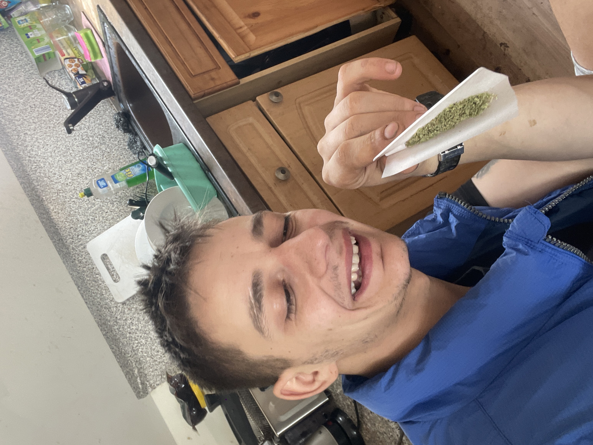 A young man with short brown hair is looking down and smiling while holding a rolled marijuana cigarette in his right hand. He is wearing a blue jacket. The background appears to be a kitchen with wooden cabinets and a sink. The scene is indoors, likely in a residential setting.