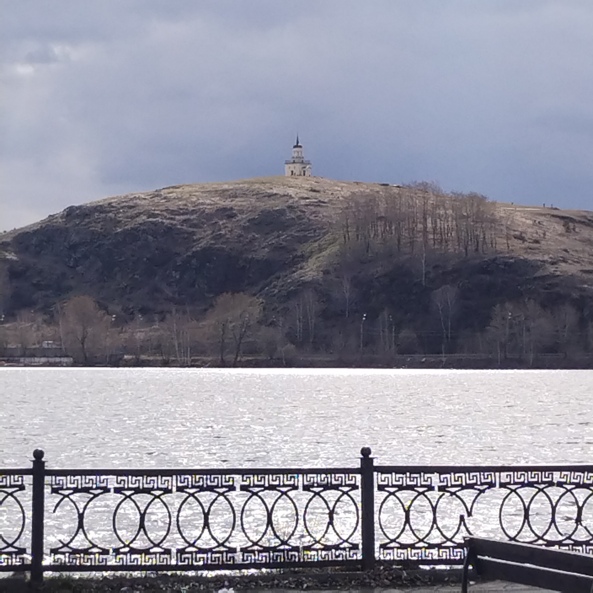 Here is a description of the image:
The image depicts a landscape scene featuring a body of water in the foreground, a hill in the midground, and a structure atop the hill. In the foreground, there is a decorative metal fence with circular and rectangular patterns and a hint of a bench, suggesting a public area or waterfront promenade. The water is reflective and appears choppy.
The midground is dominated by a hill with sparse vegetation and trees. The hill appears rugged and possibly rocky. The structure on the hill is a tower or small building with a spire. The sky is overcast, indicating a cloudy day.
The image is of the Lysaya Mountain in Nizhny Tagil, Russia, with the observation tower on top.