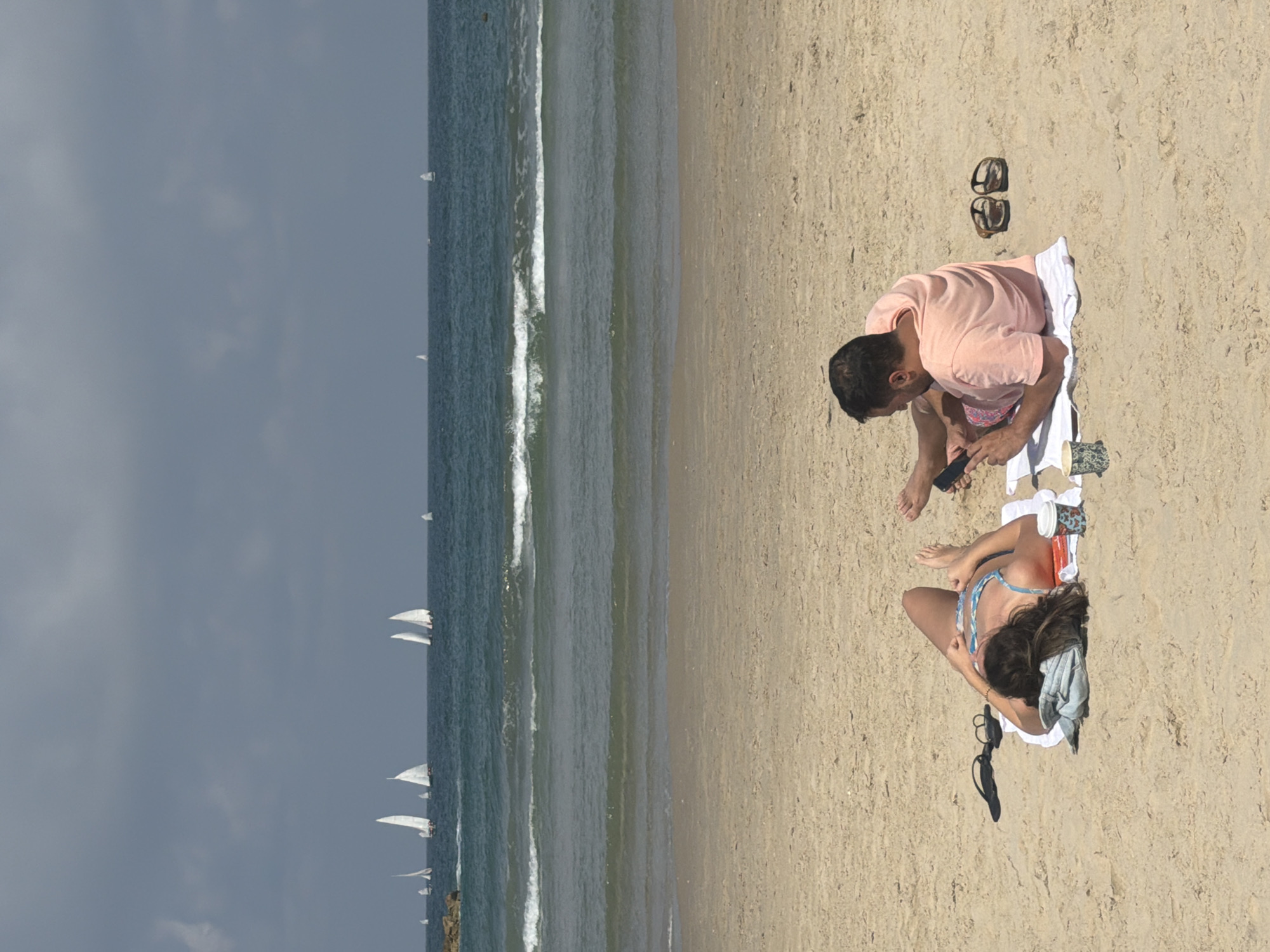 A man and a woman are sitting on a beach towel on the sand at a beach in Tel-Aviv, Israel. The man is sitting with his legs crossed and looking down at a cell phone he is holding. The woman is sitting with her legs stretched out and is turned toward the man. The sea is to the left of the image with sailboats visible in the distance.