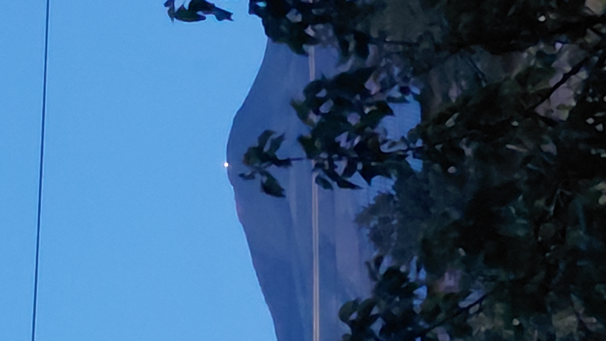 The image captures a twilight scene with a prominent mountain in the background, possibly Mount Ara, located near Abovyan, Armenia. A bright light is visible on the summit of the mountain. In the foreground, dark silhouettes of tree branches and leaves obscure the lower portion of the image. A thin, dark horizontal line, likely a power line, stretches across the upper part of the sky. The sky is a clear, pale blue, indicating it is either dusk or dawn. The overall impression is one of a natural landscape at the edge of twilight, with a man-made element (the light on the mountain) drawing attention.
