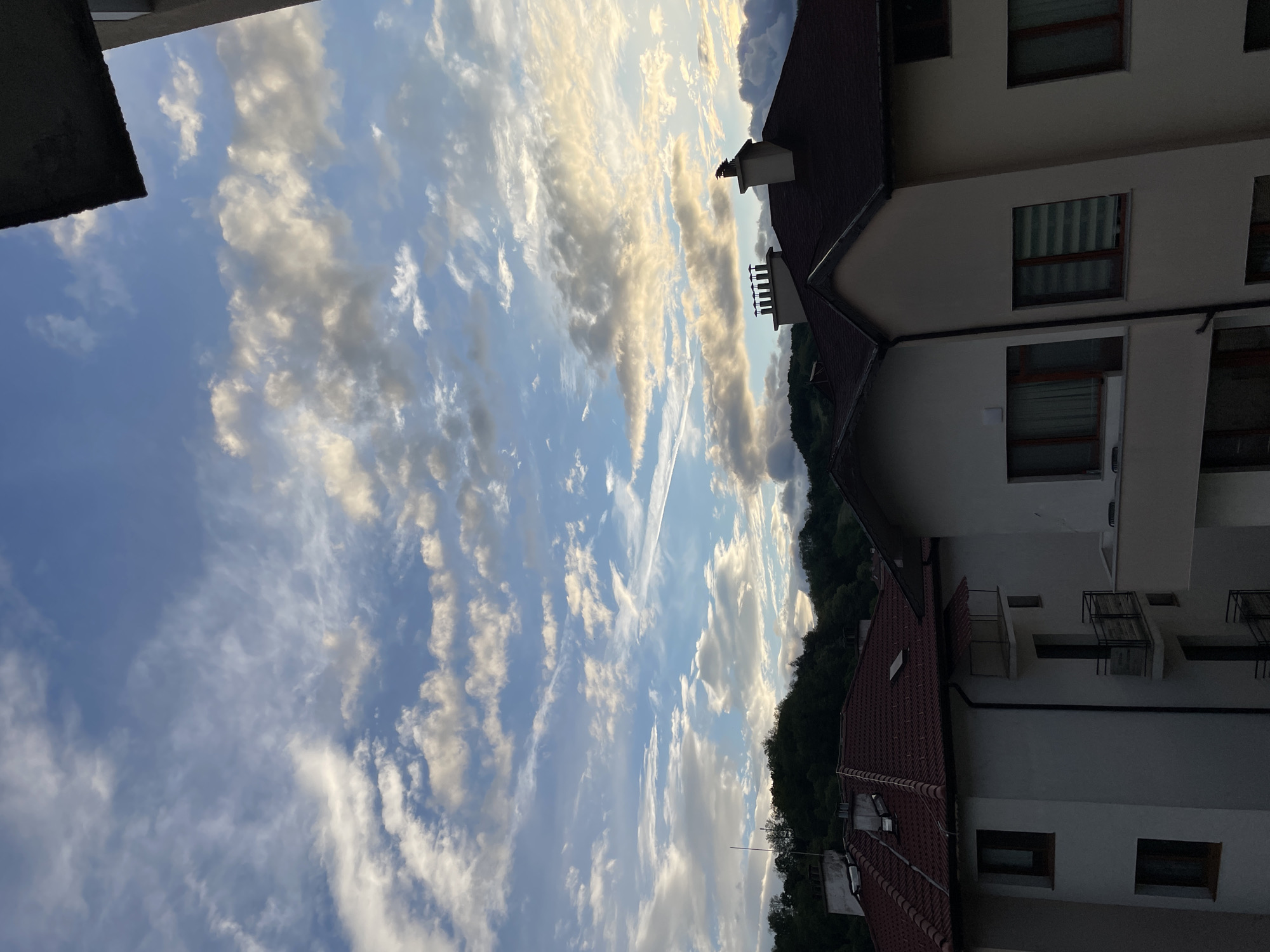 The image presents a daytime view of Zlatograd, Bulgaria, featuring a complex layering of environmental and architectural elements. Predominantly, the frame is occupied by a blue sky populated with white and grey clouds that exhibit a striated pattern. This cloudscape serves as a backdrop to the upper portions of buildings.
The buildings are multi-storied and appear to be constructed of light-colored materials with some sections clad in white. These buildings showcase elements like dark-colored roofing, windows with some featuring blinds, and balconies. The arrangement suggests they are part of a residential or mixed-use area.
Scattered trees and dense greenery are visible, suggesting the location's proximity to natural landscapes. This greenery contrasts with the built environment, adding depth and context to the scene.