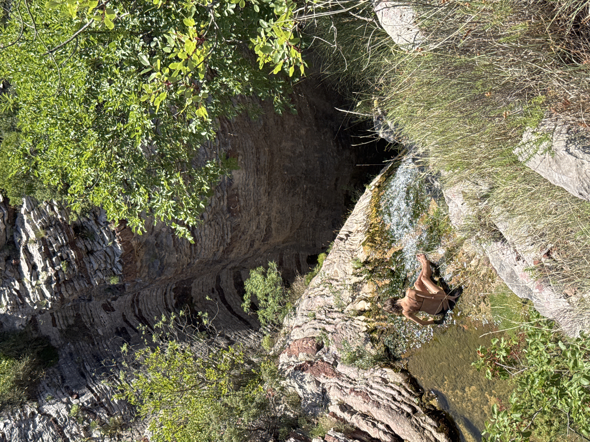 A lone person in a black bikini sits on a rocky outcrop with a waterfall cascading over the edge into a clear pool of water. The person is seen from behind, with their back to the camera. The surrounding environment is rugged and natural, with steep, layered rock formations and lush green vegetation. The scene appears to be a secluded natural spot. This location is in Rafailovići, Montenegro.