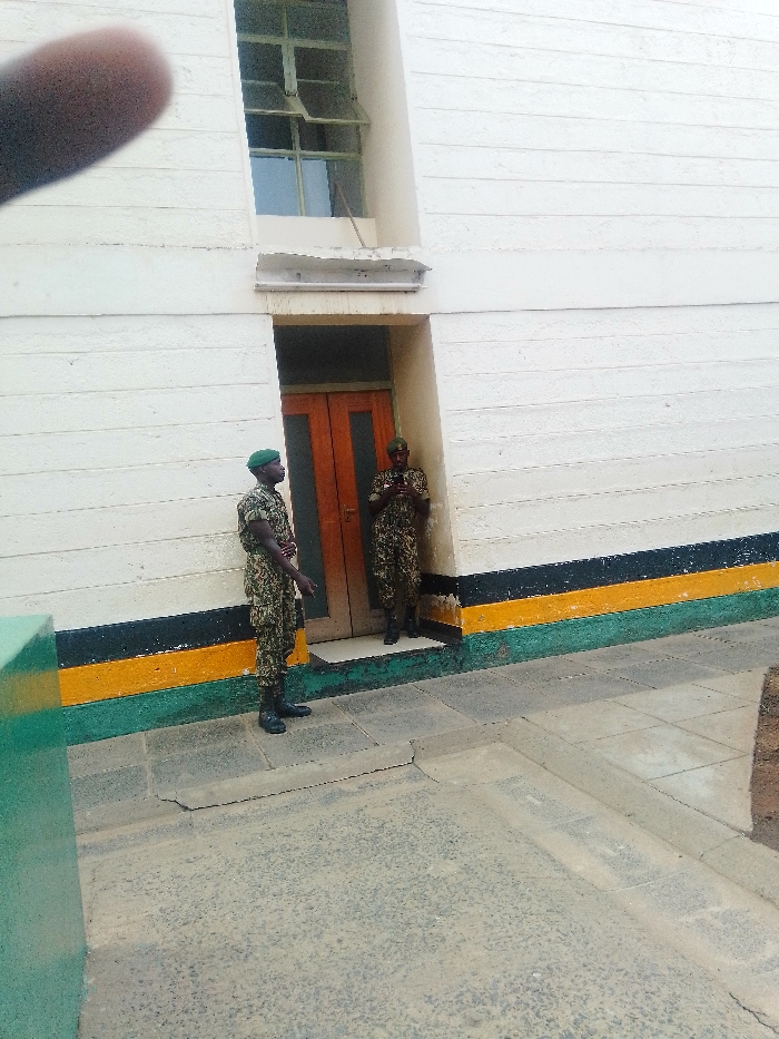 The image displays two uniformed males standing outside a building in what appears to be a government or security facility in Nairobi, Kenya. This identification is supported by the distinct horizontal black, yellow, and green stripes painted along the lower exterior wall of the building, which are the colors of the Kenyan national flag.
Both individuals are adult males dressed in full camouflage uniforms, dark boots, and green berets. The male on the left stands with his body angled slightly towards the right, arms at his sides, observing. The male on the right stands within a recessed double-leaf wooden doorway, facing slightly left, looking down at a small, dark object held with both hands, consistent with a mobile device.
The building wall is cream or off-white with horizontal textured lines. Above the doorway, a recessed window with metal frames and multiple panes has its two upper sections open outwards. The ground in the immediate foreground consists of light grey paving stones, transitioning to a darker, worn asphalt or tarmac surface further out, separated by a low curb. A partial green rectangular structure is visible in the far left foreground. A blurred human finger is present in the top-left corner of the image.