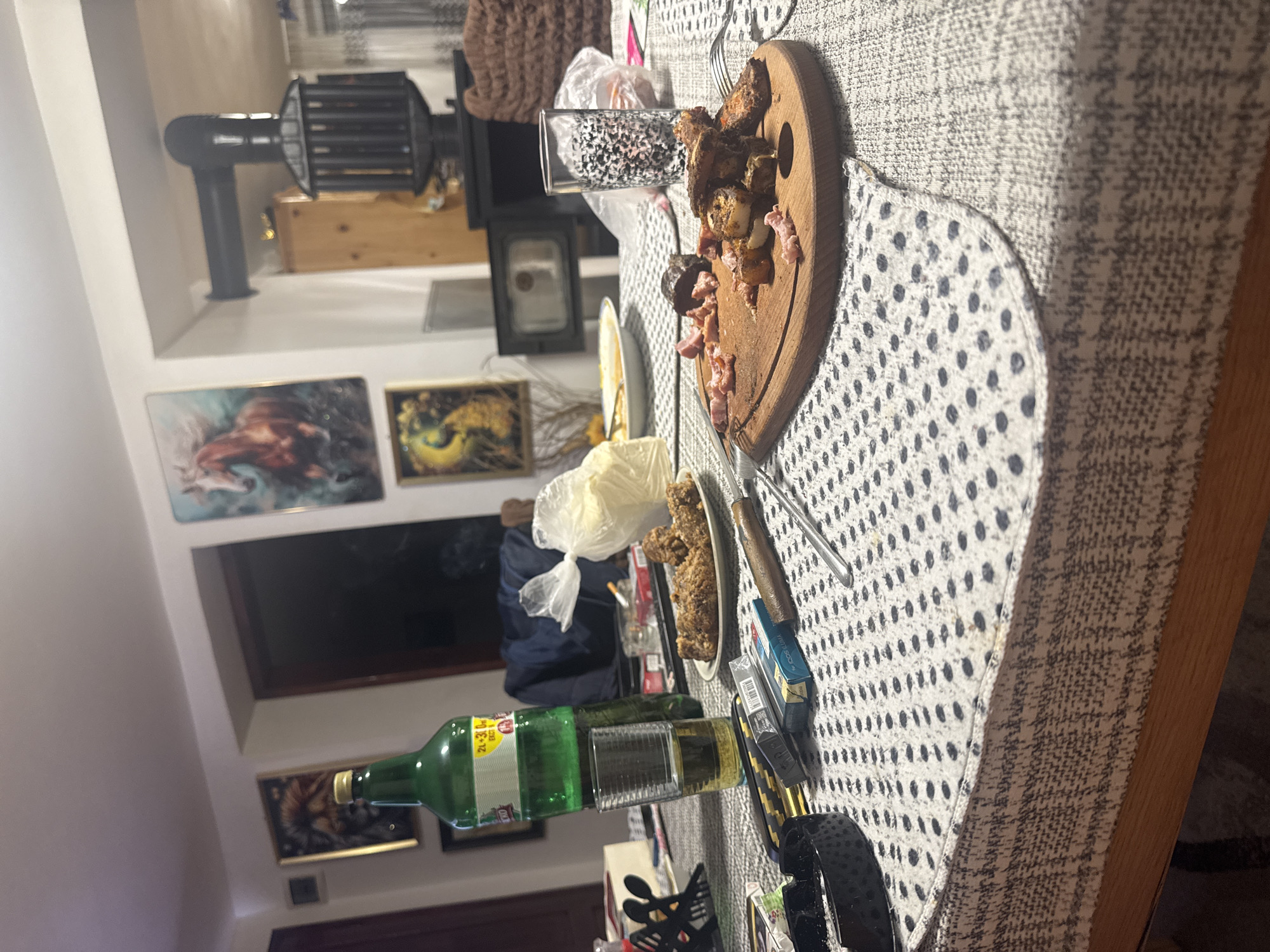 The image depicts a table laden with food and beverages. A wooden cutting board with sliced meats sits near a glass filled with dark spheres. A bottle of green liquid is lying on its side next to a pair of headphones and a pack of cigarettes. Other food items are present on plates. The table is covered with a white and grey patterned tablecloth. The background shows a room interior with paintings on the wall, a window, and a wood-burning stove. The location is Livada, Bulgaria.