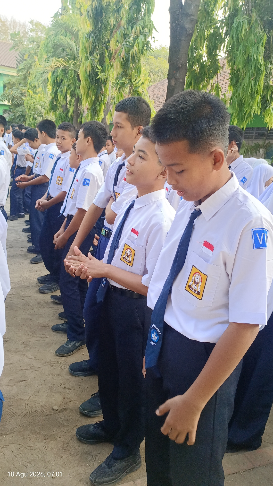This is a medium shot, eye-level, outdoor photograph taken in Kecamatan Pati, Indonesia. The image shows a group of male students standing in a line outdoors. They are all wearing white shirts, navy blue pants, and navy blue ties. Many of the students have patches on their shirts, including a red and white flag and a yellow crest with an emblem. Some of the students are also wearing blue square patches with the letter "VI" on their shirts. The students appear to be engaged in a formal activity, such as a morning assembly or flag ceremony, as they are standing at attention with their hands clasped in front of them. The background consists of trees and some buildings, suggesting the students are on a school campus. The lighting indicates it is daytime, likely morning, given the timestamp at the bottom left of the image: "18 Agu 2026, 07:01". The ground is a dusty, sandy surface. The students' faces show varying expressions, some looking forward, others slightly down. The overall scene conveys a sense of order and discipline.
