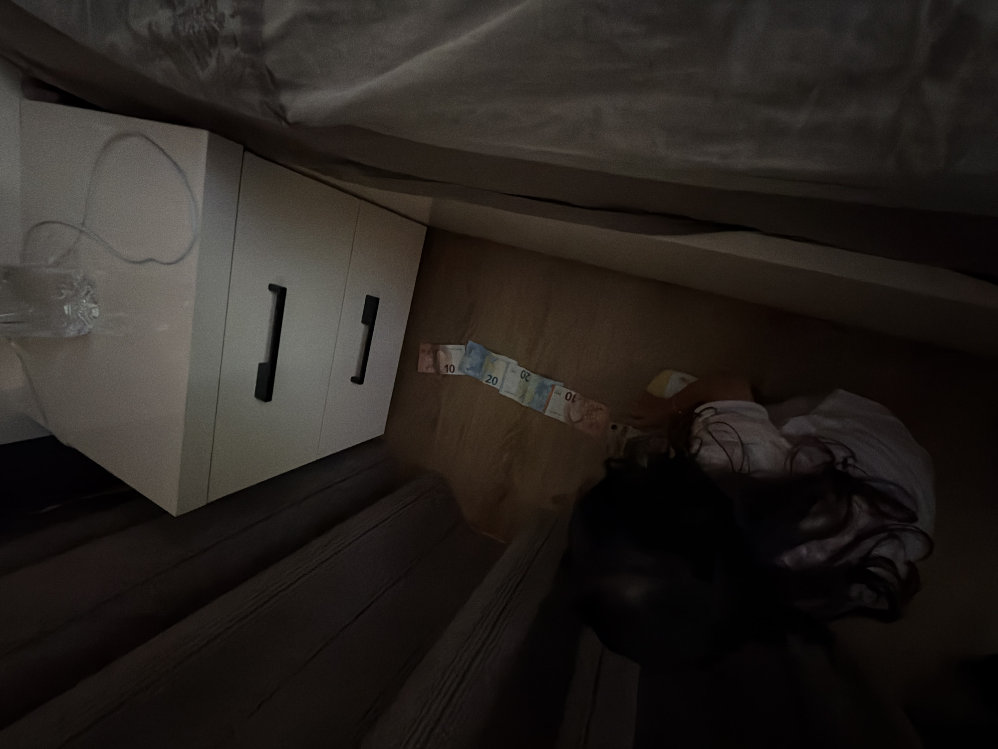 A young child, with long dark hair, is sitting on a wooden floor next to a bedside table with three drawers. The child is wearing a white t-shirt and is looking down. In front of the child, laid out on the floor, are several Euro banknotes, including 10, 20, and 50 Euro bills, arranged in a line leading away from the child towards the bedside table. The bedside table is white with black handles on each drawer. A partially filled water bottle and a charging cable are on top of the bedside table. The scene appears to be indoors, in a bedroom, with dark curtains on the left side of the image and a bed on the right. The location of Burgas, Bulgaria is not identifiable from the visual information in the image.