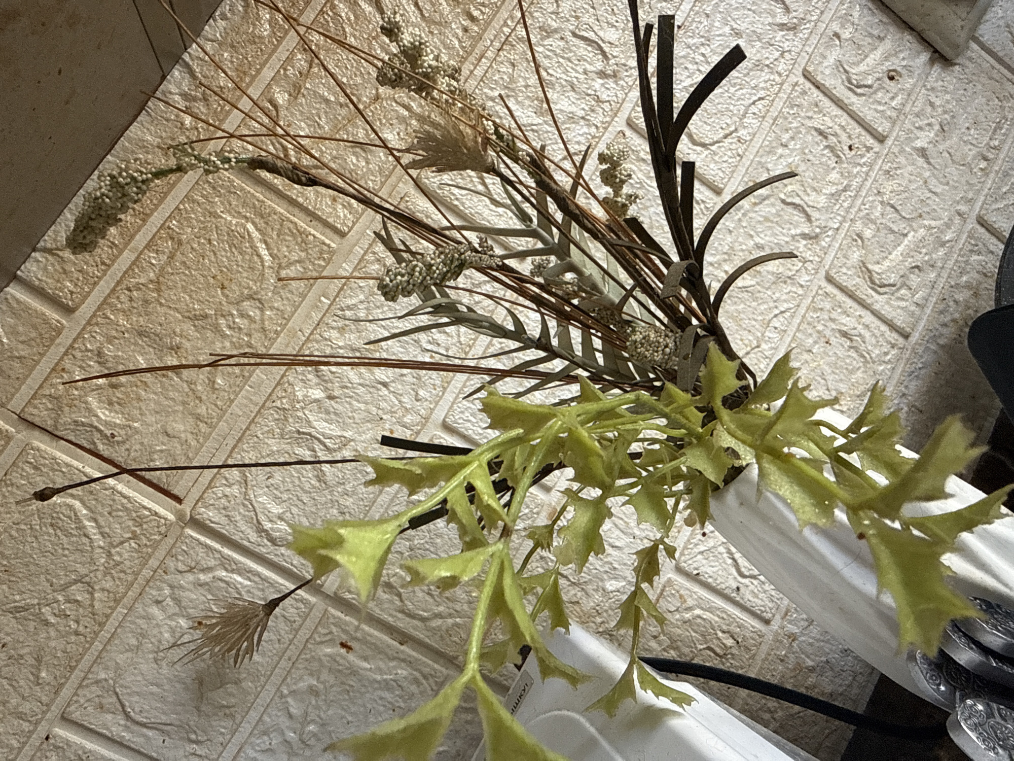 A still life image features a white ceramic vase with an arrangement of artificial plants and dried grasses. The vase is positioned in the lower left of the frame, partially obscured by the leaves of a bright green artificial plant. Behind the arrangement, a textured white wall resembling brickwork serves as the background. The lighting suggests an indoor setting, casting shadows that add depth to the composition. No people or animals are visible in the image. The location is not specified within the image content.