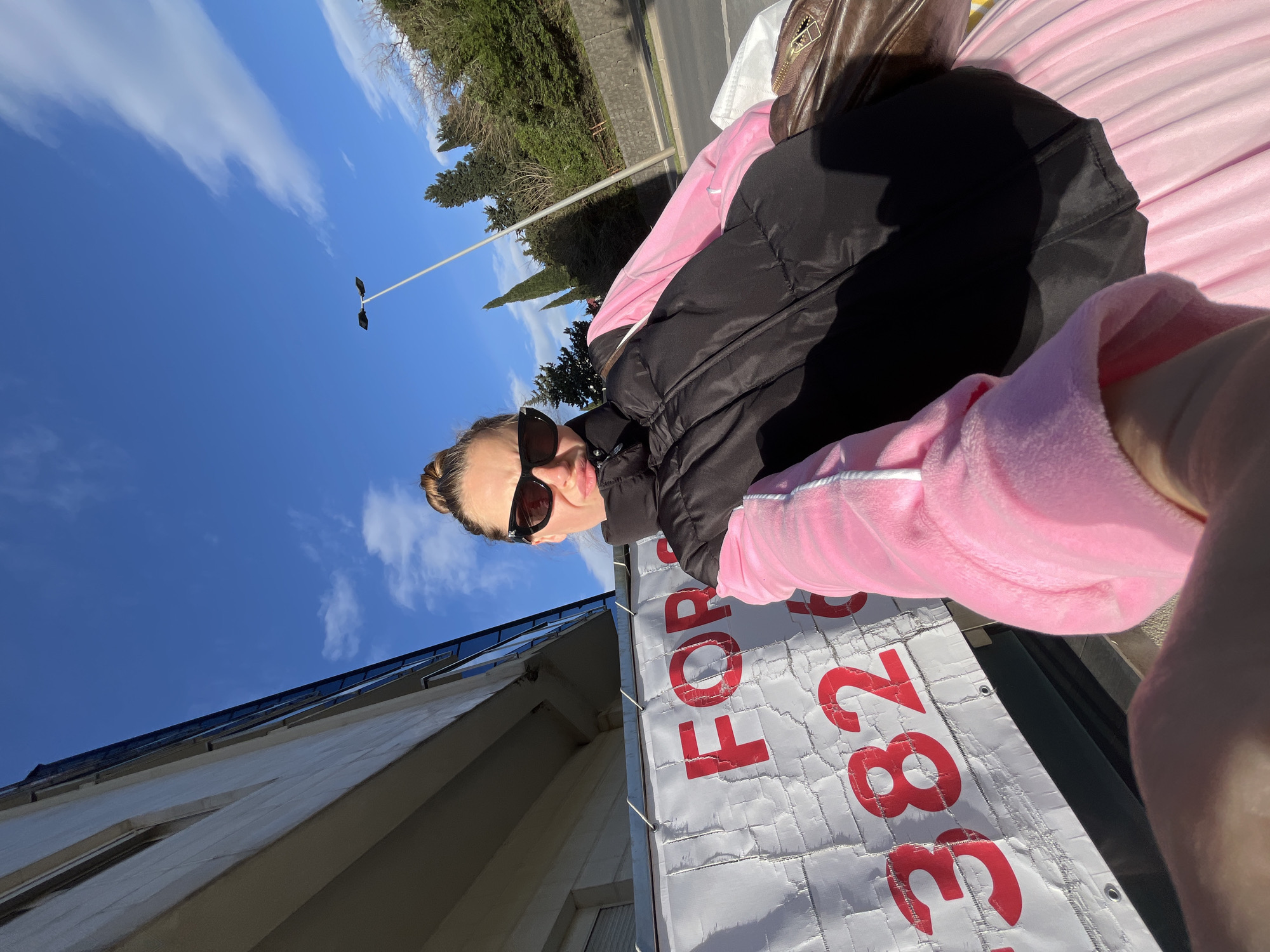 A woman wearing sunglasses, a black jacket, and a pink tracksuit is holding up a "FOR SALE" sign with the number "382" visible on it. She is standing near a building, and there is a street with trees in the background. The sky is blue with some clouds. The image appears to have been taken in Boreti, Montenegro, based on the context.