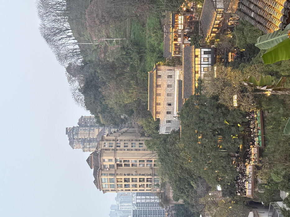The image depicts a hilly urban landscape at twilight in Nan'an District, China. The scene is dominated by a mix of modern high-rise residential buildings and traditional Chinese architecture set against a densely vegetated hillside.
In the background, on the left, numerous modern high-rise residential towers, light in color and multi-storied, rise towards an overcast, pale grey sky. One prominent, beige-colored high-rise with a complex roofline is visible in the mid-ground left, partially integrated into the hillside. To the right, a steep, heavily wooded hill is covered in various trees, some bare, suggesting deciduous species, and others evergreen. Utility poles and lines are discernible on this slope.
In the mid-ground center and right, a cluster of traditional Chinese-style buildings is nestled into the hillside. A two-story grey stone building with a prominent traditional tiled roof, illuminated with warm yellow light, stands out. Below and to its right, multiple smaller commercial structures with dark wooden frames, light-colored walls, and traditional dark tiled roofs are visible, their eaves and signs (e.g., "SHIBA BAR," "EAT'S BAR") brightly illuminated with warm yellow lights.
The foreground on the left and center features several elevated outdoor platforms built into the hillside. These platforms are densely populated with hundreds of individuals, visible as small figures, gathered at tables or standing. The areas are brightly lit by string lights and other fixtures, indicating a lively social or dining environment. Large, mature trees with dense green foliage are integrated into these platforms. In the lower right foreground, a section of a traditional tiled roof is visible, along with broad green leaves, possibly from banana plants.