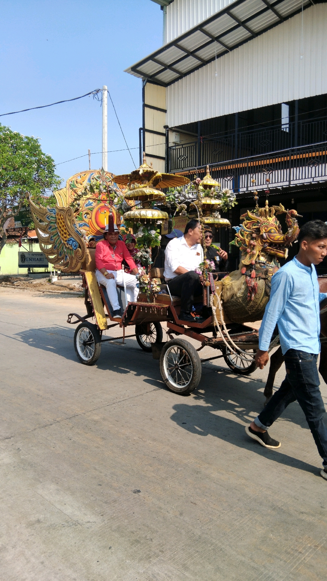 In Batang, Indonesia, on a sunny day with a clear blue sky, a horse-drawn carriage, elaborately decorated with golden dragon motifs, flowers, and ceremonial umbrellas, travels down a paved road. The carriage is occupied by three men. The driver, a man wearing a red shirt, white pants, and a hat, sits at the front. Behind him, two other men are seated, one in a white shirt and dark pants, and another in a darker outfit. The horse pulling the carriage is visible, and a young man in a blue shirt and dark pants walks alongside it, also appearing to be part of the procession. The background features a modern building with a white corrugated metal facade and dark railings, suggesting an urban or commercial setting. A tree with green foliage is also visible to the left of the carriage. The overall scene depicts a cultural or festive event.
