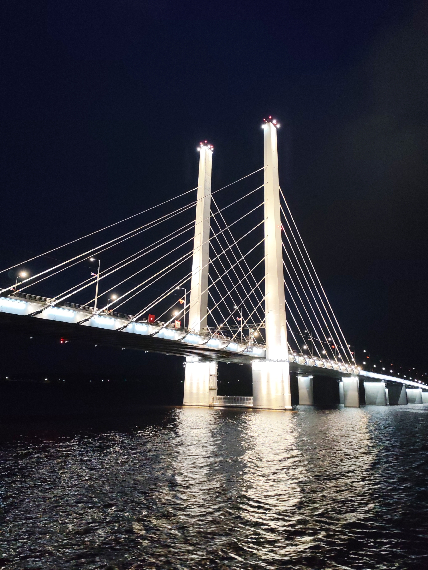 This image shows a cable-stayed bridge at night. The bridge features two tall, white towers with lights at the top and a series of cables connecting the towers to the bridge deck. The deck is lit with streetlights and has small red and white flags. The bridge spans a body of water, with light reflecting off the water's surface. The sky is dark. The location is a city that is Unknown, Unknown.