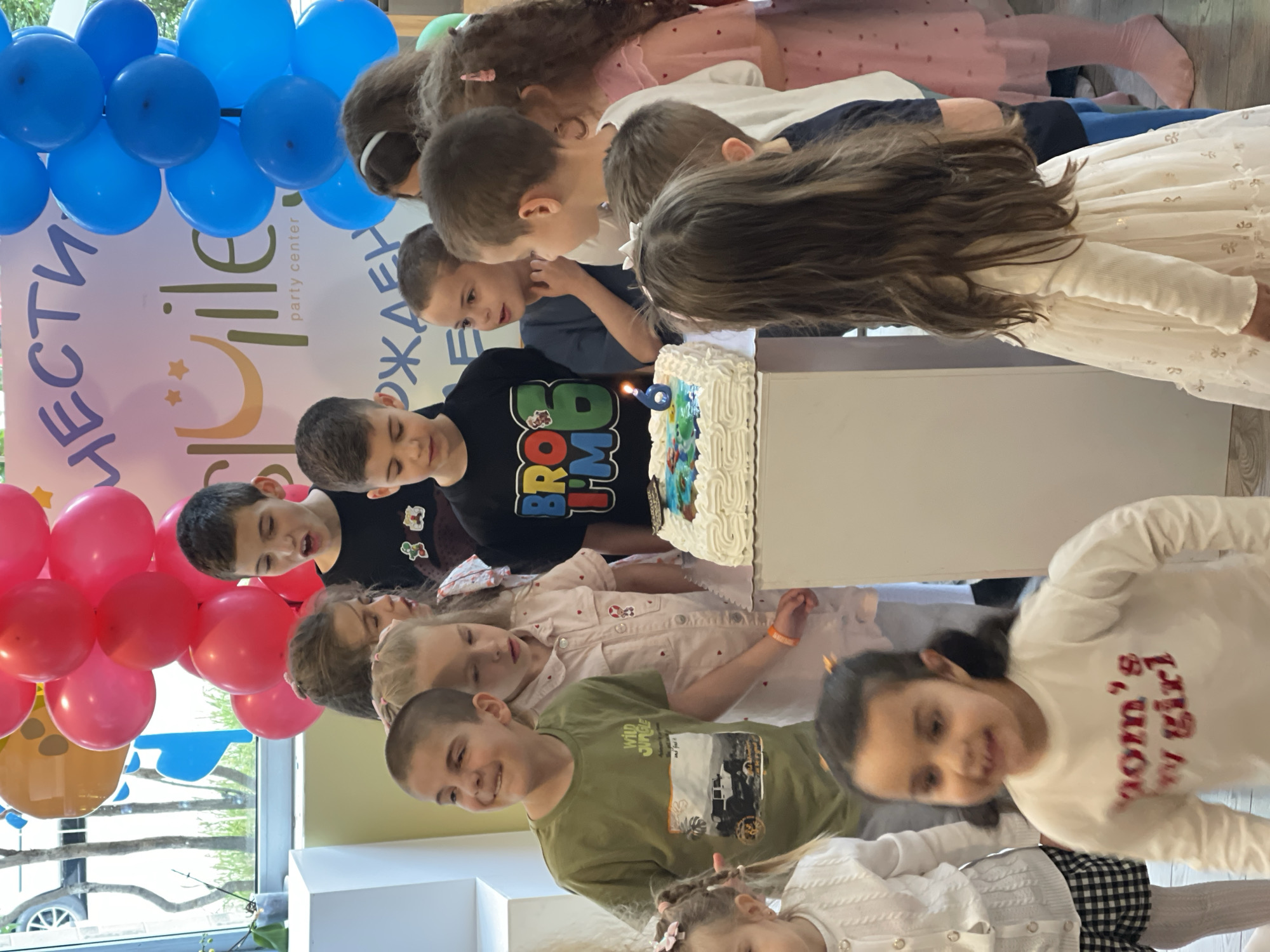 A group of children is gathered around a birthday cake with a number '6' candle, likely at a party. The event seems to be taking place at "Ville party center" (as indicated by the text on the balloon arch), which is located in Sofia, Bulgaria. The children are of varying ages and are looking at the cake with anticipation. There are blue and pink balloons decorating the scene. The children are wearing casual clothing.