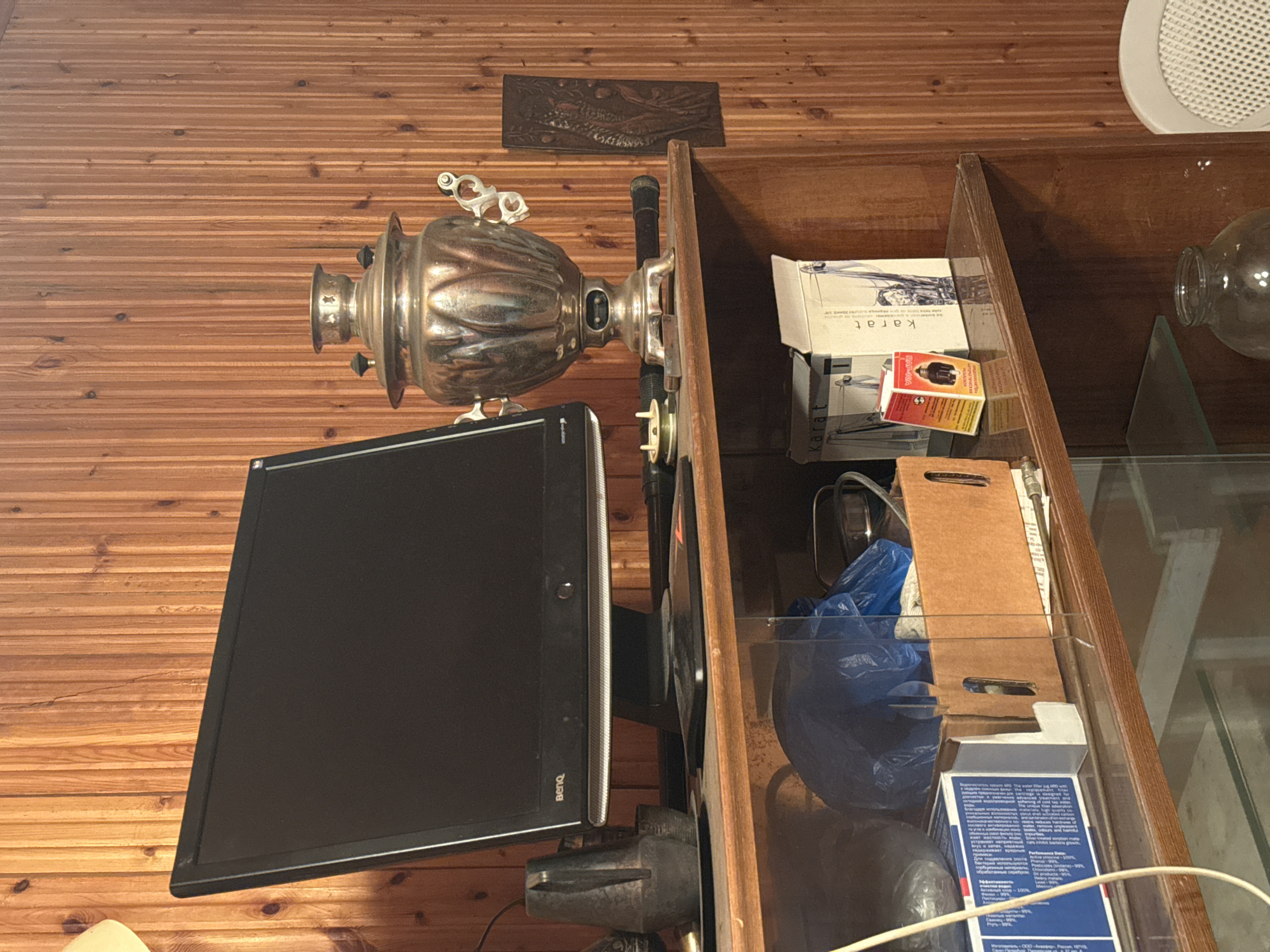 The image shows a room with a wood-paneled wall and a glass-shelved cabinet. A samovar sits near a black computer monitor. Inside the cabinet are various items including boxes (one labeled "karat"), a blue plastic bag, and a glass bottle.