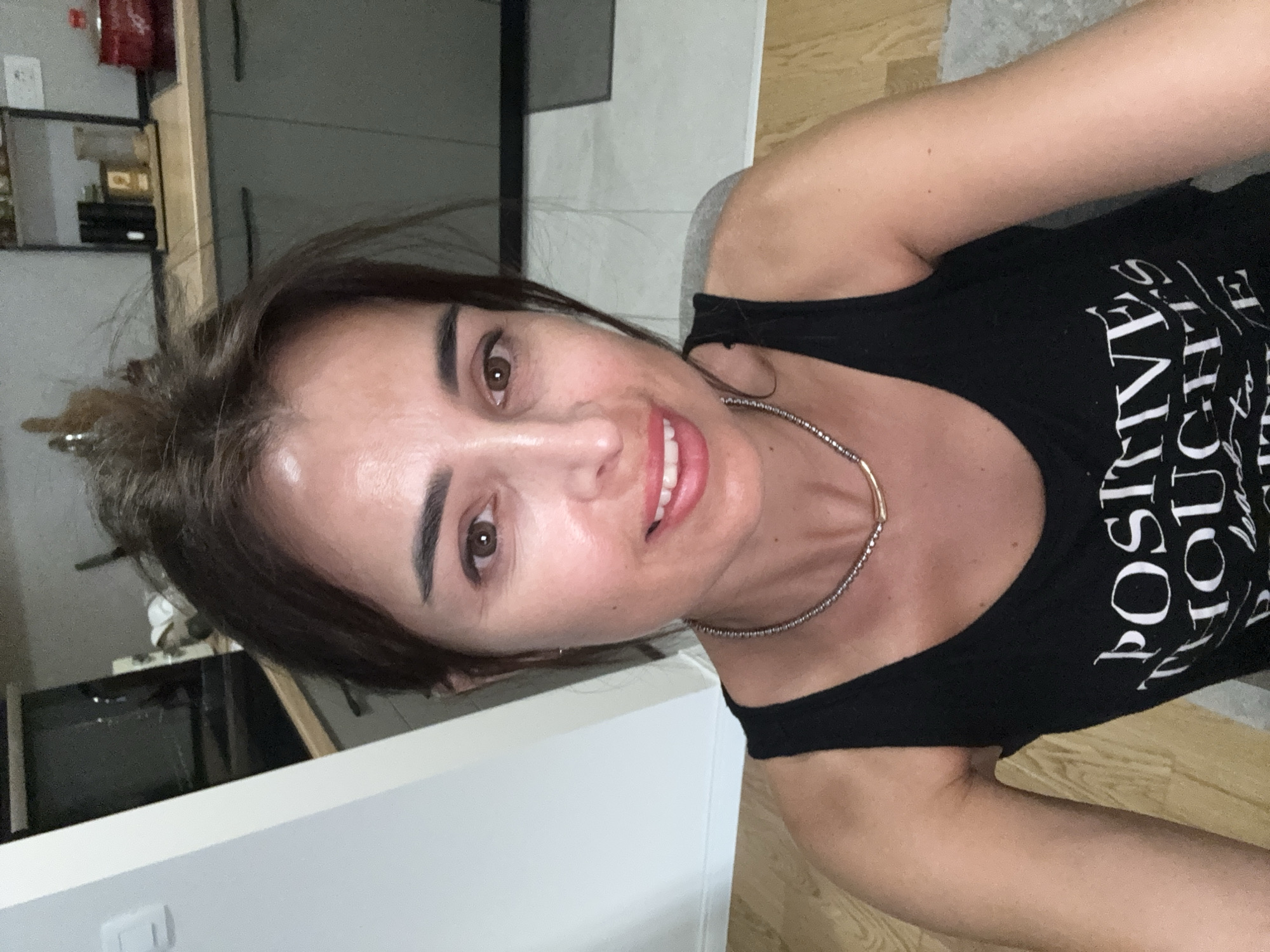 A woman with brown hair, pulled up into a messy bun, is looking at the camera. She is wearing a black tank top with white text that reads "POSITIVE THOUGHTS LEAD TO..." and a silver necklace with gold accents. She is smiling and her eyes are open. The background appears to be a kitchen with gray cabinets and a wooden countertop. There are shelves with various items on them. The lighting is dim. The woman is positioned in the foreground, taking up most of the frame.