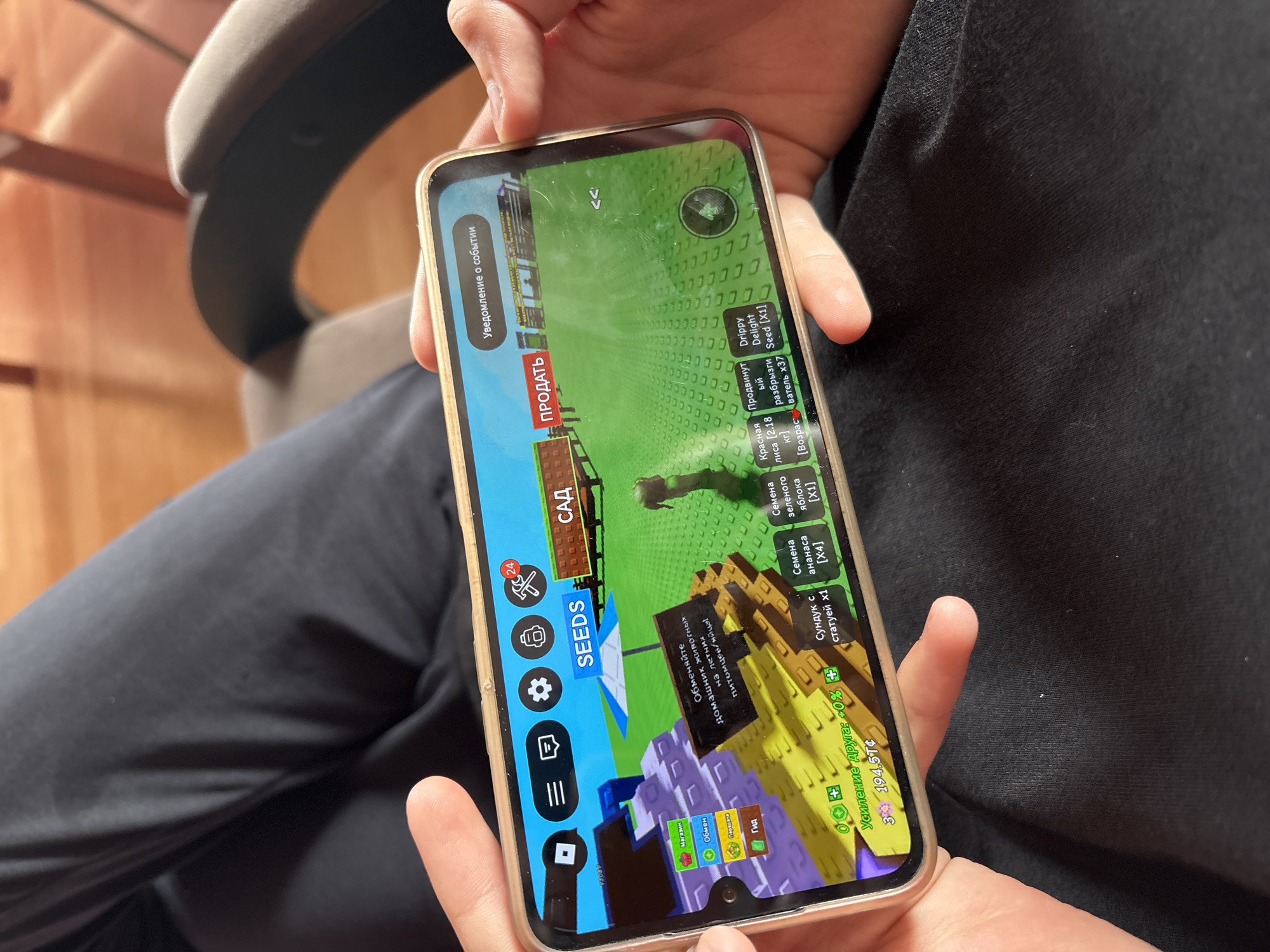 The image shows a person holding a smartphone in landscape mode, displaying a game interface. The game appears to be a farming or simulation game with colorful 3D graphics. The interface includes buttons for "SEEDS" and "ПРОДАТЬ" (Sell). There are also icons for settings, inventory, and notifications. In the foreground, a finger is visible interacting with the screen. The background shows a person's legs wearing black pants. The environment outside the phone's display is out of focus, with hints of furniture and a wooden floor, suggesting an indoor setting. There is no indication of the city of Ветлужский, Russia, in the image.