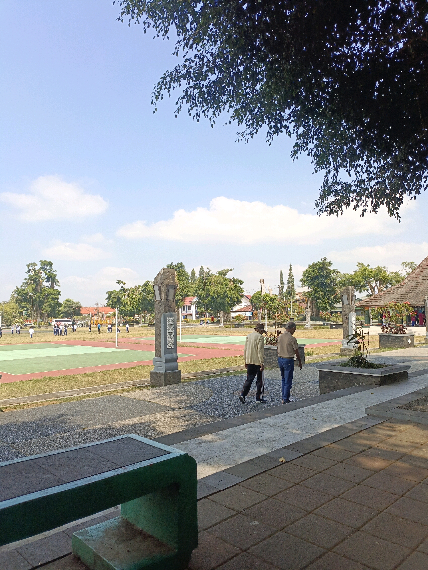 This is a medium-angle, full-shot, vertical photograph taken outdoors on a bright, sunny day in the city of Wonosobo, Indonesia, likely at Alun-Alun Wonosobo, a public square. In the foreground, a green-painted, concrete bench with a dark gray tiled seat is positioned at a slight angle to the camera, its left side facing the viewer. The bench is situated on a paved walkway made of dark gray square tiles.
To the right of the bench, the walkway continues, bordered by a raised rectangular planter box containing a sparse, thorny plant. Beyond this, a second walkway, made of lighter gray and white rectangular tiles, leads further into the scene. Two elderly men are walking away from the camera on this lighter walkway. The man on the left wears a light tan long-sleeved shirt and dark pants with a white stripe down the side. The man on the right wears a light brown short-sleeved shirt and blue jeans.
In the middle ground, a large open space features several sports courts. One court, closest to the viewer, has green and red sections, possibly for volleyball or badminton, with a net visible. Further back, other courts are visible, with people, possibly students in uniforms, scattered around them. A bus and other vehicles are parked in the distance. Several trees, some large and mature, dot the landscape, and buildings with terracotta roofs are visible behind the trees. A stone monument, resembling a lamp post base with carvings, stands to the right of the sports courts, between the foreground and middle ground. The sky is a clear, light blue with scattered white clouds, and the branches of a large tree with dark green leaves overhang the top right portion of the image, casting dappled shadows. The scene conveys a sense of everyday activity in a public recreational space.