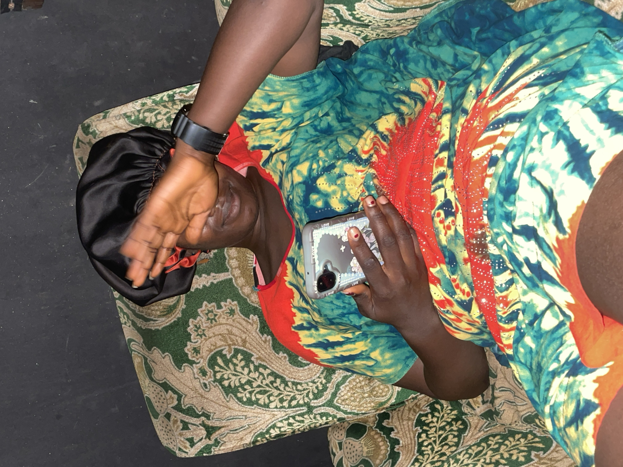 A person wearing a black bonnet and a colorful tie-dye dress with red, yellow, and blue patterns sits on a green and beige patterned armchair. They are holding a smartphone in their right hand and their left hand is covering their face. A black watch is visible on their left wrist. The background appears to be a dark, textured wall. The image captures a domestic setting, likely within a home in Jos, Nigeria, given the prompt's context.