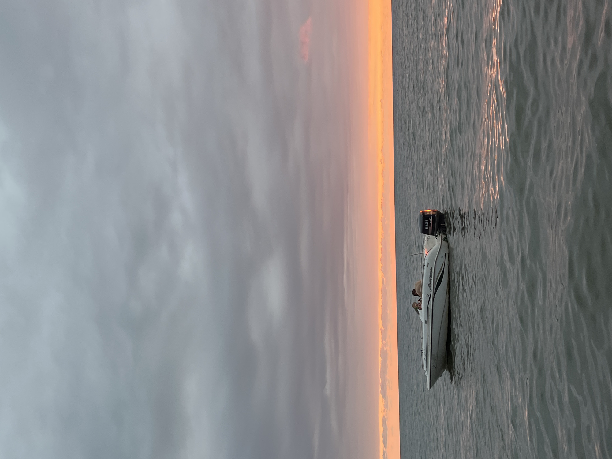 An outdoor, low-angle, medium shot captures a boat with two people aboard on a calm body of water. The boat, white with a dark stripe, is positioned in the center of the frame. Two individuals, one appearing to be an adult and the other a child, are seated within the boat. The boat has a Yamaha outboard motor attached to its stern.
The environment is a large expanse of water under a cloudy sky. The horizon is visible, separating the water from the sky. The sky exhibits a gradient of colors, with darker gray clouds at the top transitioning to a soft orange and pink hue near the horizon, suggesting either sunrise or sunset. The water is a muted gray, reflecting the sky, with subtle ripples and the glint of sunlight on its surface. The scene is devoid of any urban structures or specific landmarks identifiable as being within the city of Ureki, Georgia, based solely on the visual information provided.