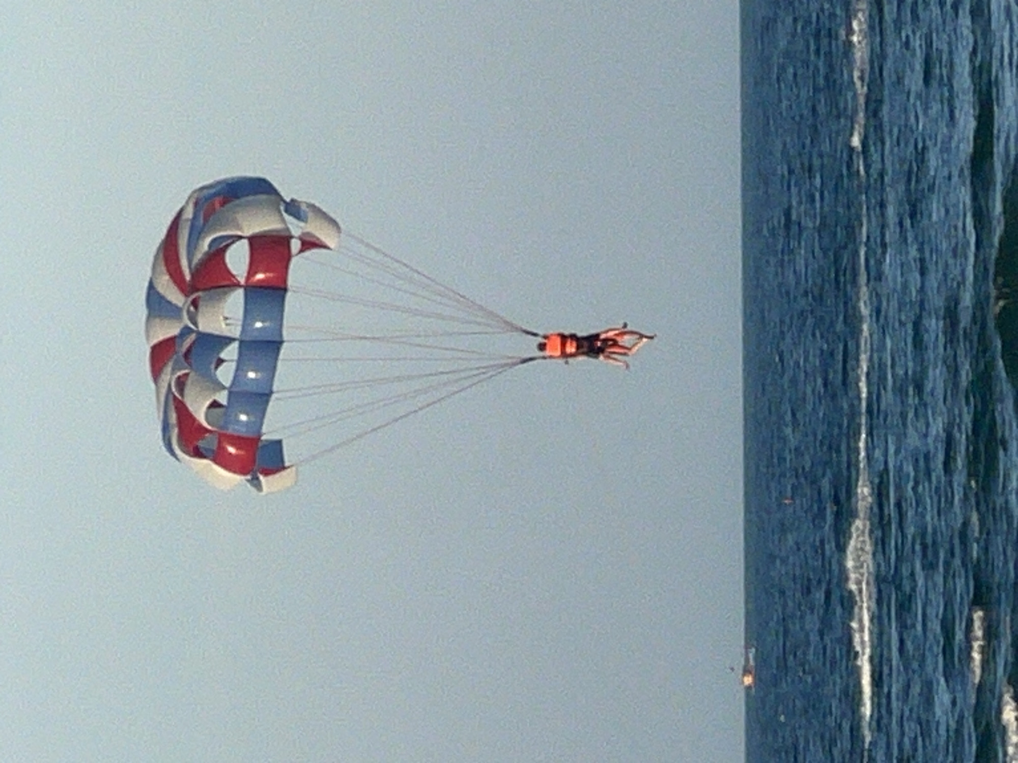 A wide shot shows a parasailer being towed behind a boat over the ocean. The parasail is red, white, and blue. Two people are harnessed to the parasail, one in front of the other. The ocean is blue with whitecaps. A small boat is visible on the horizon. The sky is clear and blue. The scene is likely Da Nang, Vietnam, a popular tourist destination known for its beaches and water sports.