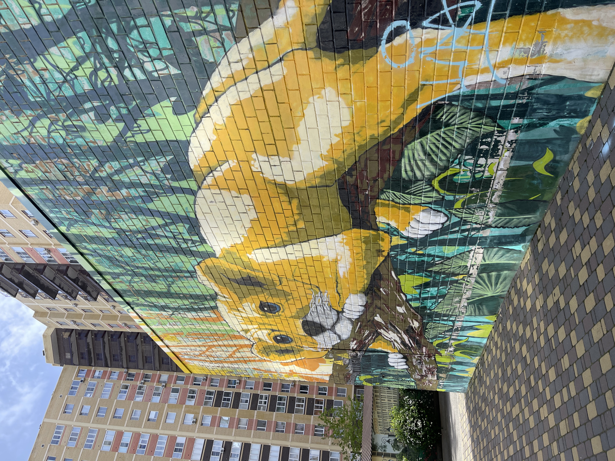A large mural depicting a yellow animal with white markings (possibly a lion cub) is painted on a brick wall in what appears to be Stavropol, Russia. The mural includes greenery, possibly representing foliage or jungle. The wall abuts a paved walkway made of yellow and gray paving stones. A tall apartment building with many windows and balconies is visible to the left of the mural, and a small tree and shrubs are at the base of the wall near the walkway. The sky is visible in the top left corner of the frame.