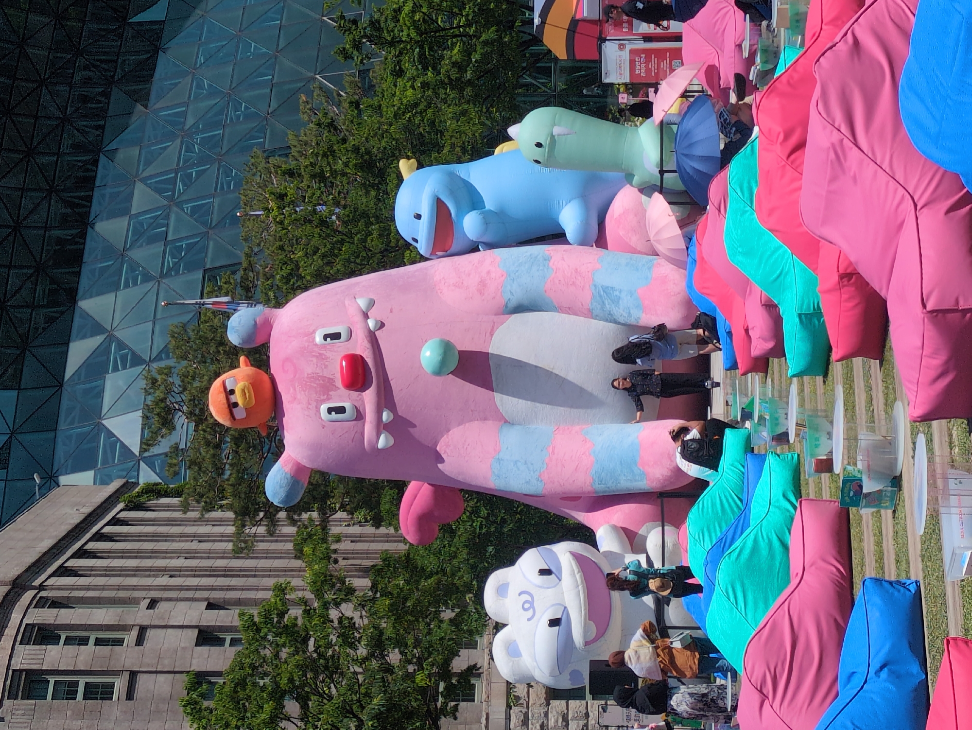 The image captures an outdoor scene in Seoul, South Korea, featuring large, inflatable cartoon characters. A pink, striped creature with an orange bird on its head dominates the foreground, along with a blue whale-like figure and a white, cloud-shaped character. People are standing near the figures, interacting with the display. In the background, there's a modern glass structure with a geometric design, contrasting with a more traditional building made of grey stone. A row of colorful beanbag chairs in pink, turquoise, and blue is set up with small, round tables and clear containers, suggesting an outdoor event or exhibition.