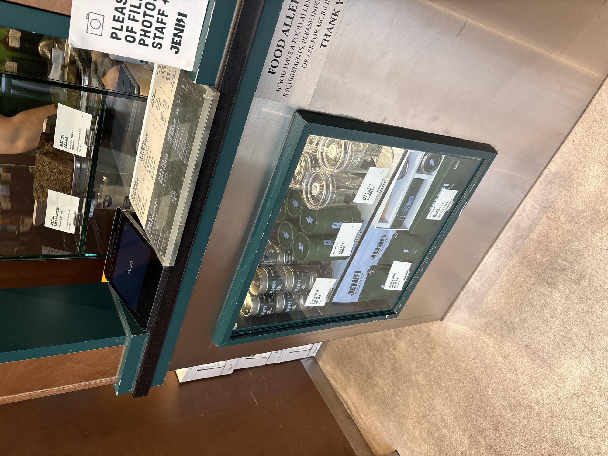 This image, captured inside a commercial establishment in London, United Kingdom, shows a cafe or matcha shop counter displaying various food and retail items. The scene is well-lit, suggesting daytime.
On the left side of the counter, a glass display case holds baked goods. Two prominent items are visible: "MATCHA BANANA BREAD WITH CHOCOLATE CHIPS" and a "MATCHA COOKIE WITH WHITE CHOCOLATE + RASPBERRY," with allergy information also listed for the cookie. An arm is partially visible behind this display, possibly a staff member's. Adjacent to the baked goods, a digital display shows "£0.00," indicating a transaction or pricing display.
To the right, a glass-encased refrigerated display cabinet showcases a variety of packaged matcha products and accessories from the brand "JENKI." Items include green tins of "CEREMONIAL GRADE MATCHA," "BAMBOO WHISK," a "JENKI ICED MATCHA TUMBLER," and boxes for "THE JENKI SET," "RITUAL STARTER SET," "DISCOVERY SET," and "HOJICHA STARTER SET."
Several signs are visible on the counter. One sign on the far left, partially obscured, appears to request "PLEASE REFRAIN OF FILMING/PHOTOS OF STAFF," mentioning "JENKI." Another prominent sign on the right side of the counter reads: "FOOD ALLERGIES: IF YOU HAVE A FOOD ALLERGY OR DIETARY REQUIREMENTS, PLEASE INFORM US OR ASK FOR MORE INFORMATION. THANK YOU." Additional menu-like signage lists categories such as "MATCHA ORIGINALS," "MATCHA SIGNATURES," "MATCHA SOFT-SERVE," and "HEALTHY BITES." The overall impression is that of a specialized matcha cafe or store.