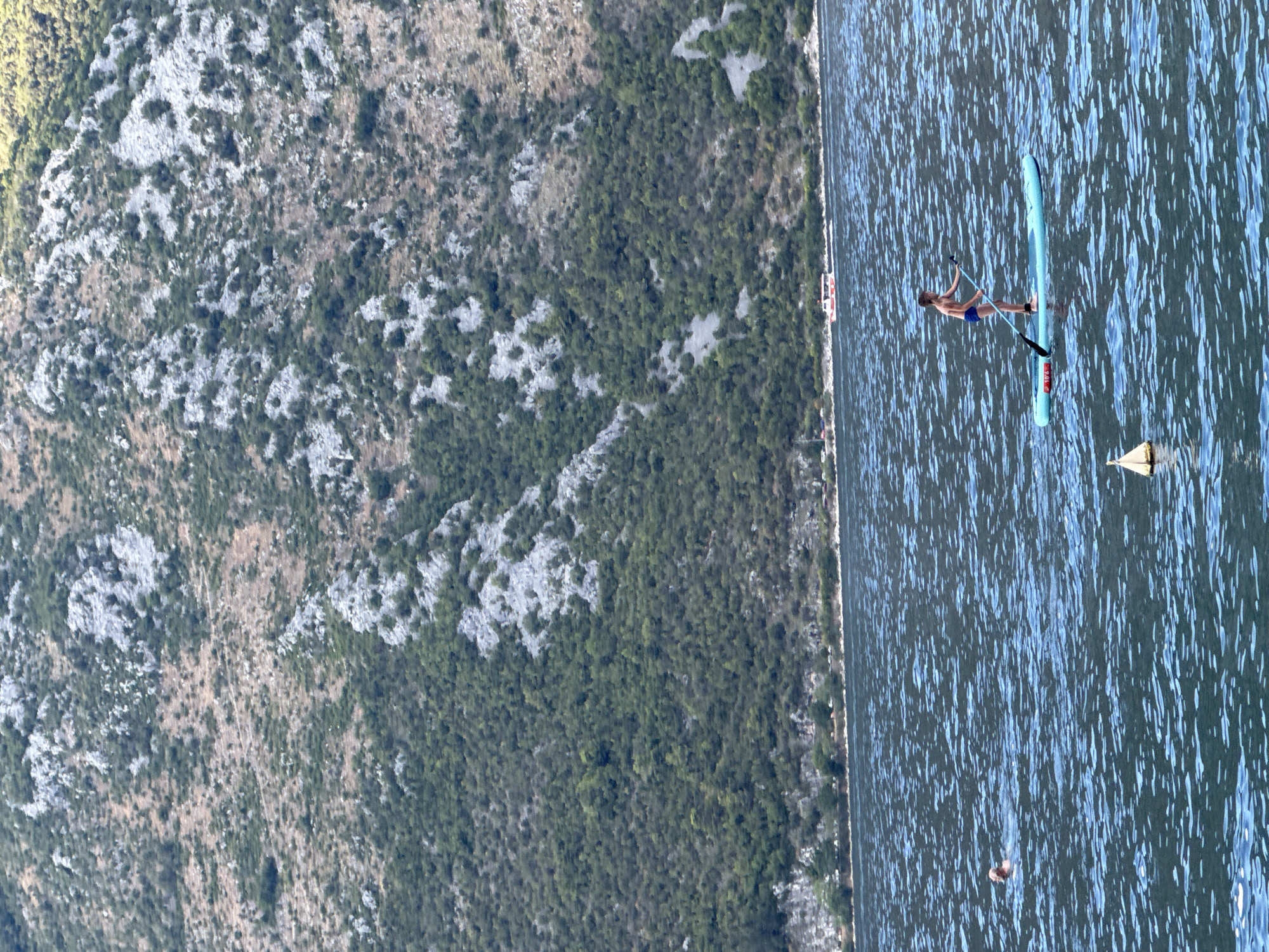 In Prčanj, Montenegro, the image captures a wide view of a bay with calm, blue water. In the foreground, a person is paddleboarding on a turquoise board. To the left, a second person is swimming in the water. In the background, a boat with red and white accents is moving across the bay. Behind the water, a steep, rocky mountain covered in sparse green vegetation rises dramatically. The shoreline at the base of the mountain features a line of trees and some structures, indicating a populated area. The overall scene is bathed in daylight.